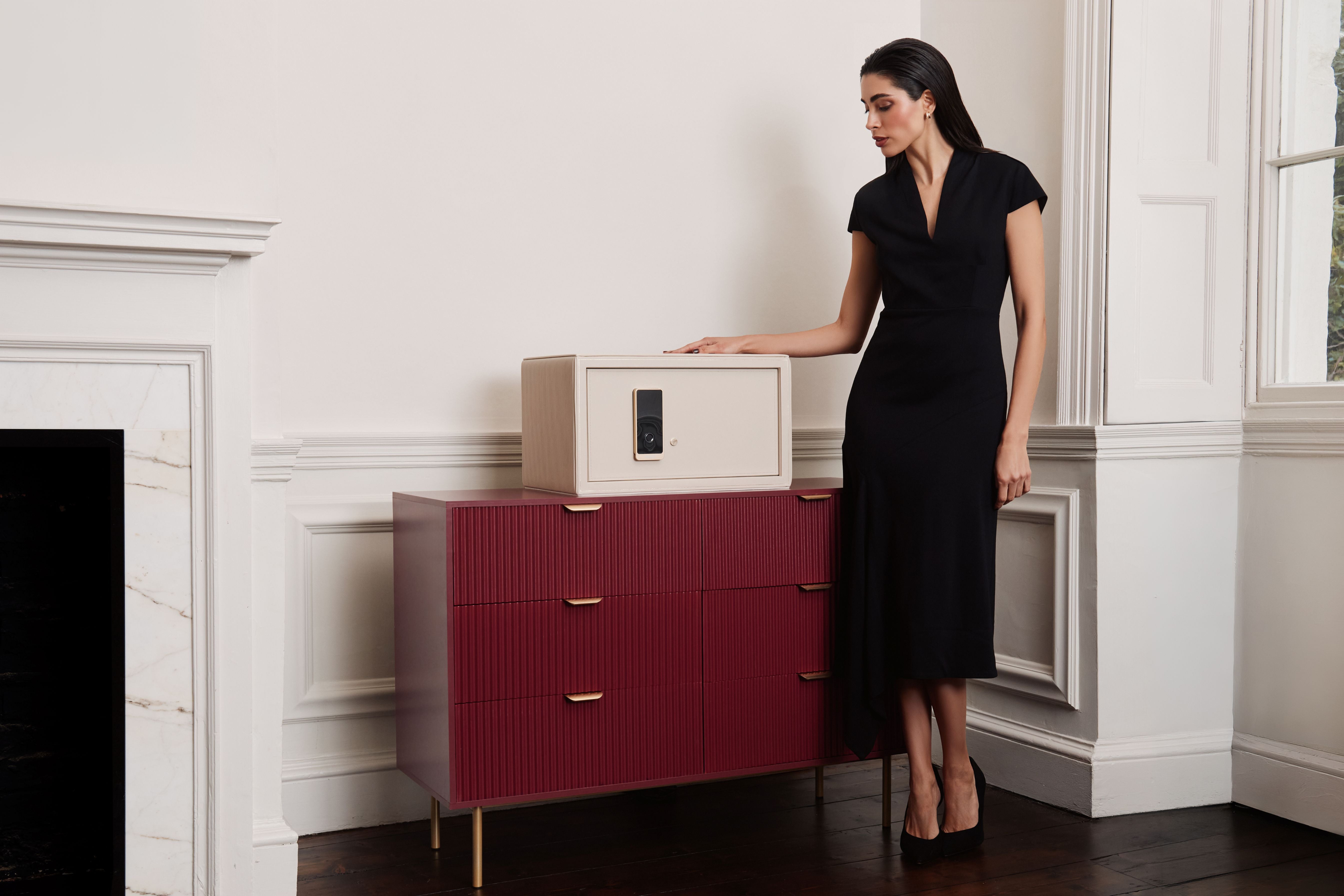 Coffre fort à bijoux Bella Den Wolf 1834 de couleur crème à ouverture biométrique. Vue lifestyle, avec le coffre fermé posé sur une commode rouge et une femme élégante habillée de noir qui se tient à coté, la main posée dessus