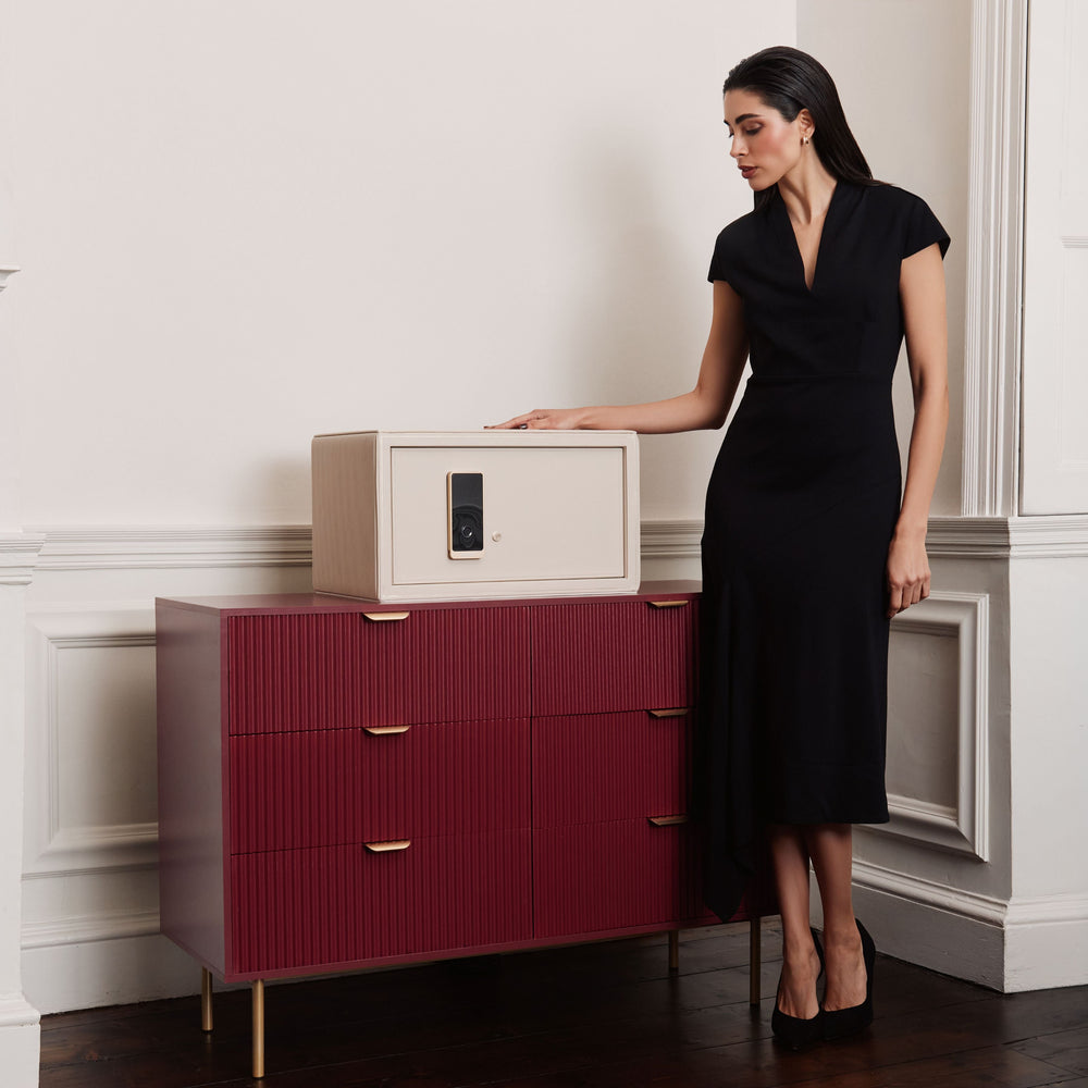 Coffre fort à bijoux Bella Den Wolf 1834 de couleur crème à ouverture biométrique. Vue lifestyle, avec le coffre fermé posé sur une commode rouge et une femme élégante habillée de noir qui se tient à coté, la main posée dessus