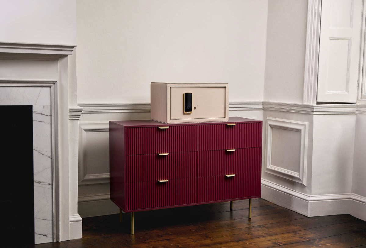 Coffre fort à bijoux Bella Den Wolf 1834 de couleur crème à ouverture biométrique. Vue lifestyle, avec le coffre fermé posé au centre d'une commode rouge 