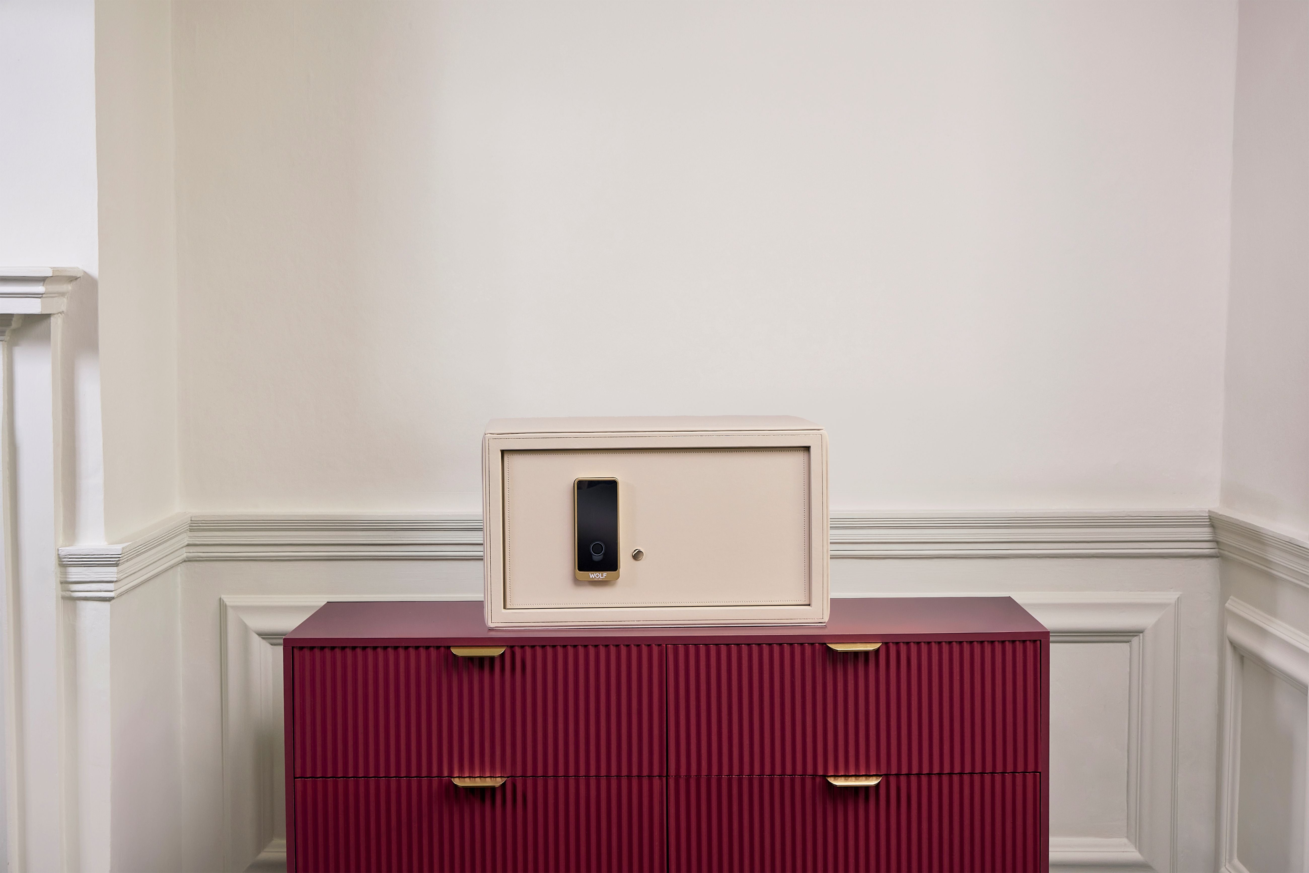 Coffre fort à bijoux Bella Den Wolf 1834 de couleur crème à ouverture biométrique. Vue lifestyle, avec le coffre fermé posé au centre d'une commode rouge 