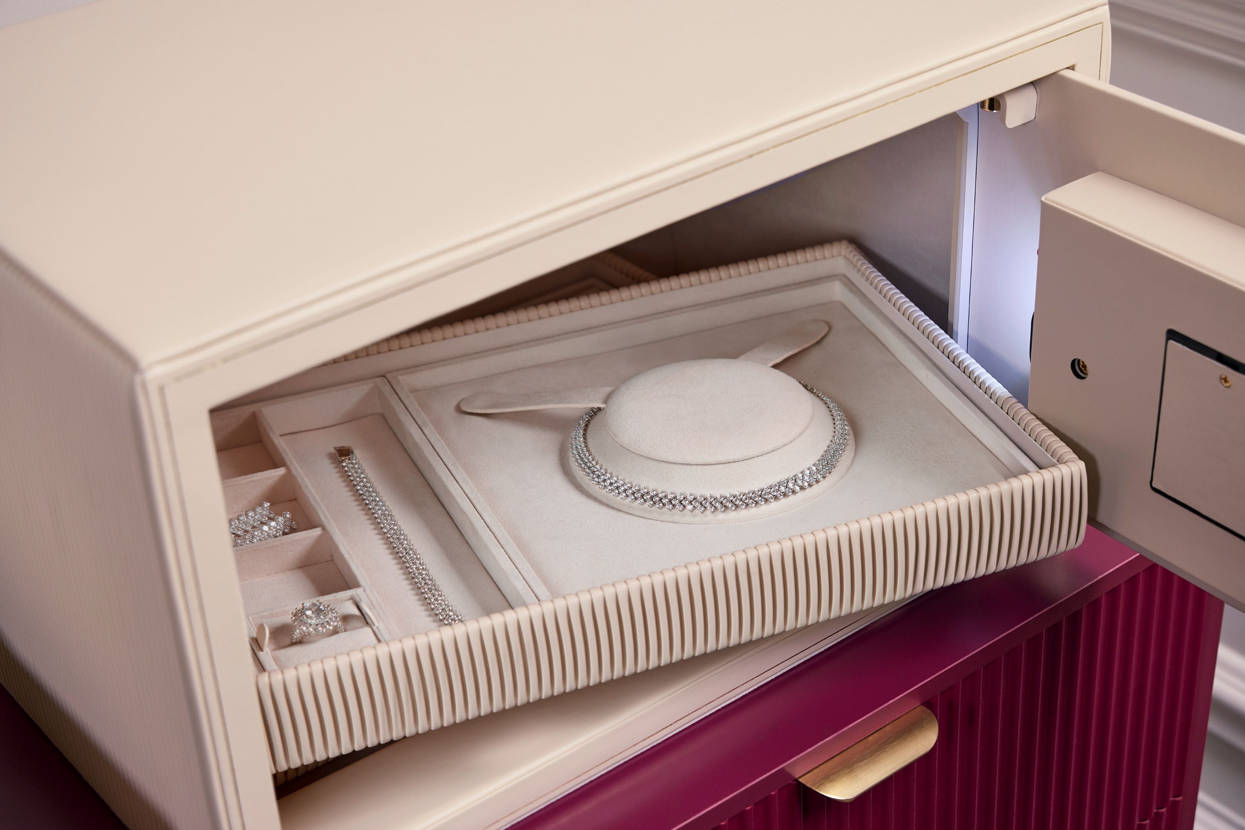 Coffre fort à bijoux Bella Den Wolf 1834 de couleur crème à ouverture biométrique. Vue lifestyle, avec le coffre ouvert posé sur une commode rouge et un zoom sur les plateaux à bijoux remplis à l'intérieur