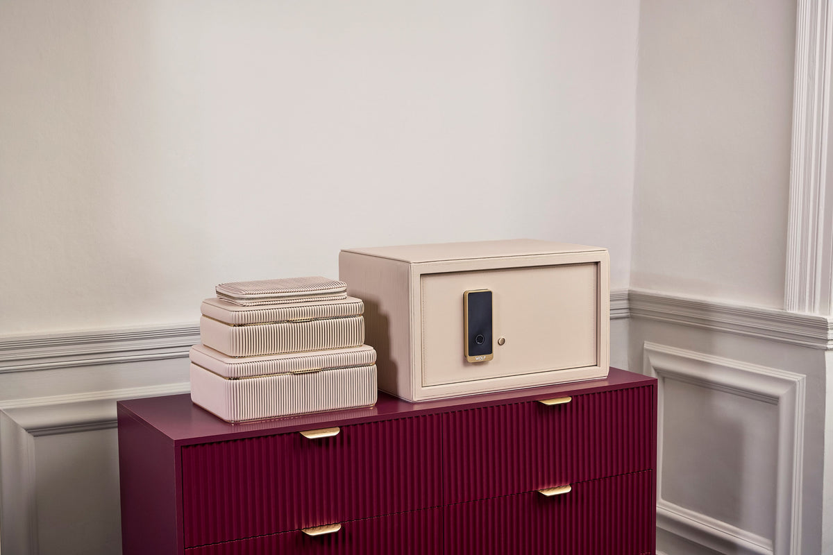 Coffre fort à bijoux Bella Den Wolf 1834 de couleur crème à ouverture biométrique. Vue lifestyle, avec le coffre ouvert posé sur une commode rouge et la collection de boîte à bijoux Bella crème posée à côté