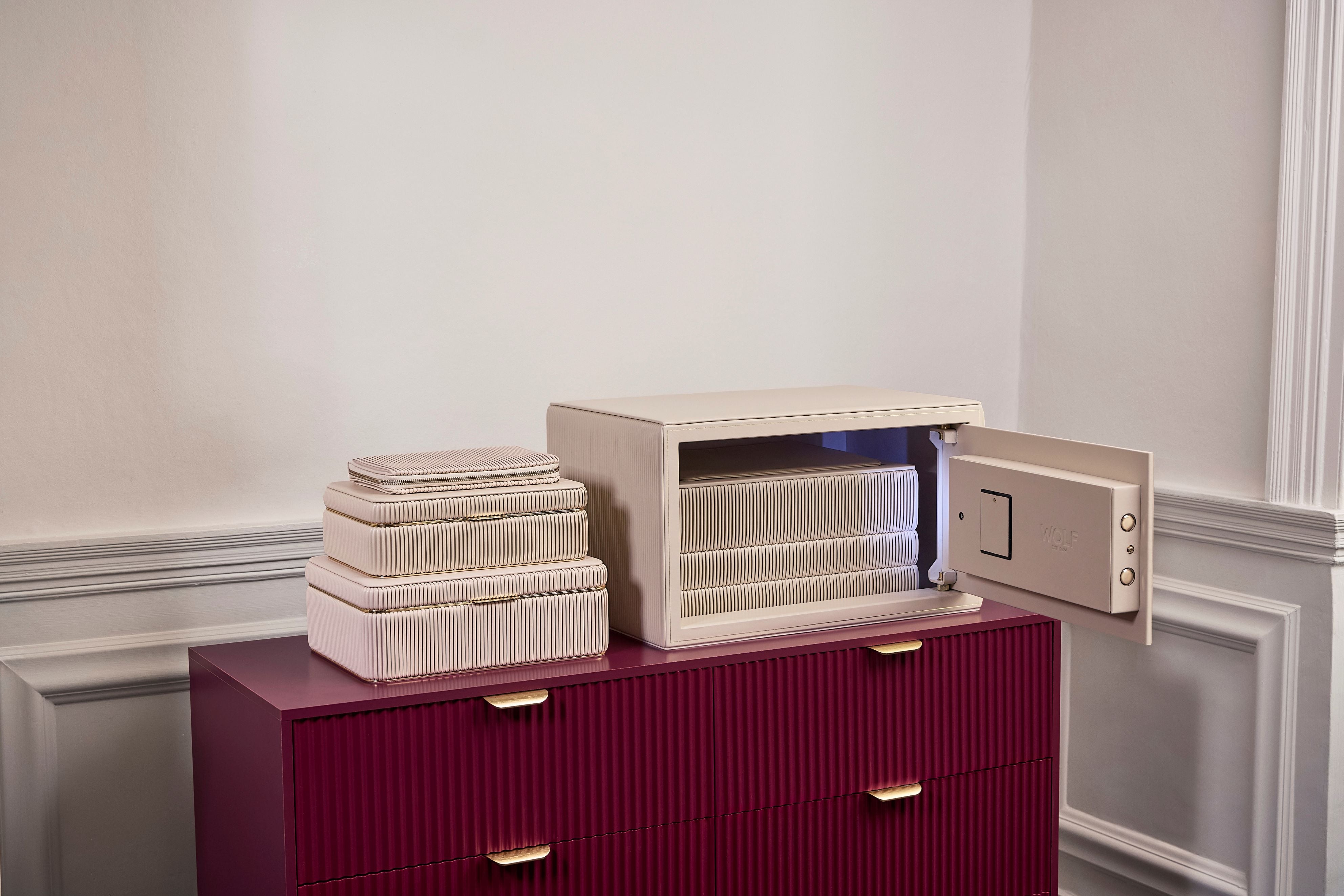 Coffre fort à bijoux Bella Den Wolf 1834 de couleur crème à ouverture biométrique. Vue lifestyle, avec le coffre ouvert posé sur une commode rouge et la collection de boîte à bijoux Bella crème posée à côté
