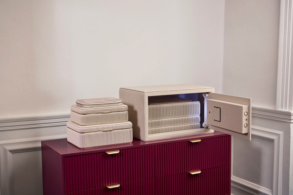 Coffre fort à bijoux Bella Den Wolf 1834 de couleur crème à ouverture biométrique. Vue lifestyle, avec le coffre ouvert posé sur une commode rouge et la collection de boîte à bijoux Bella crème posée à côté