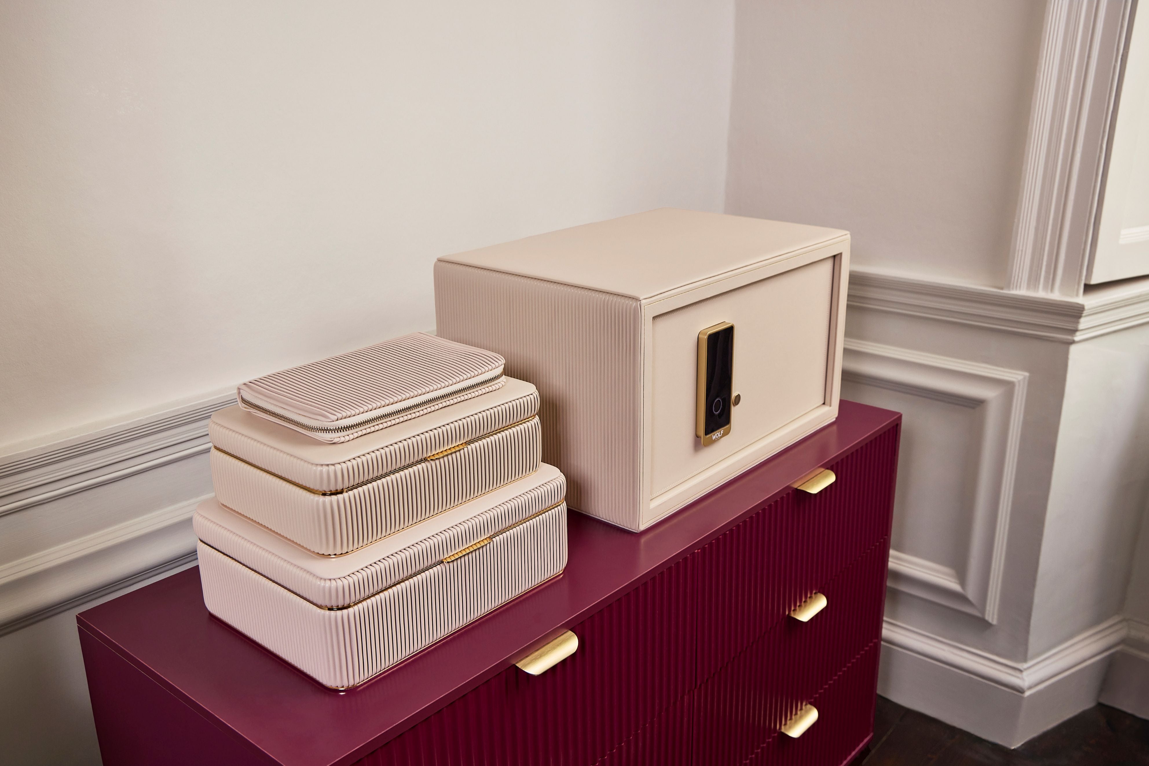 Coffre fort à bijoux Bella Den Wolf 1834 de couleur crème à ouverture biométrique. Vue lifestyle, avec le coffre fermé posé sur une commode rouge et la collection de boîte à bijoux Bella crème posée à côté