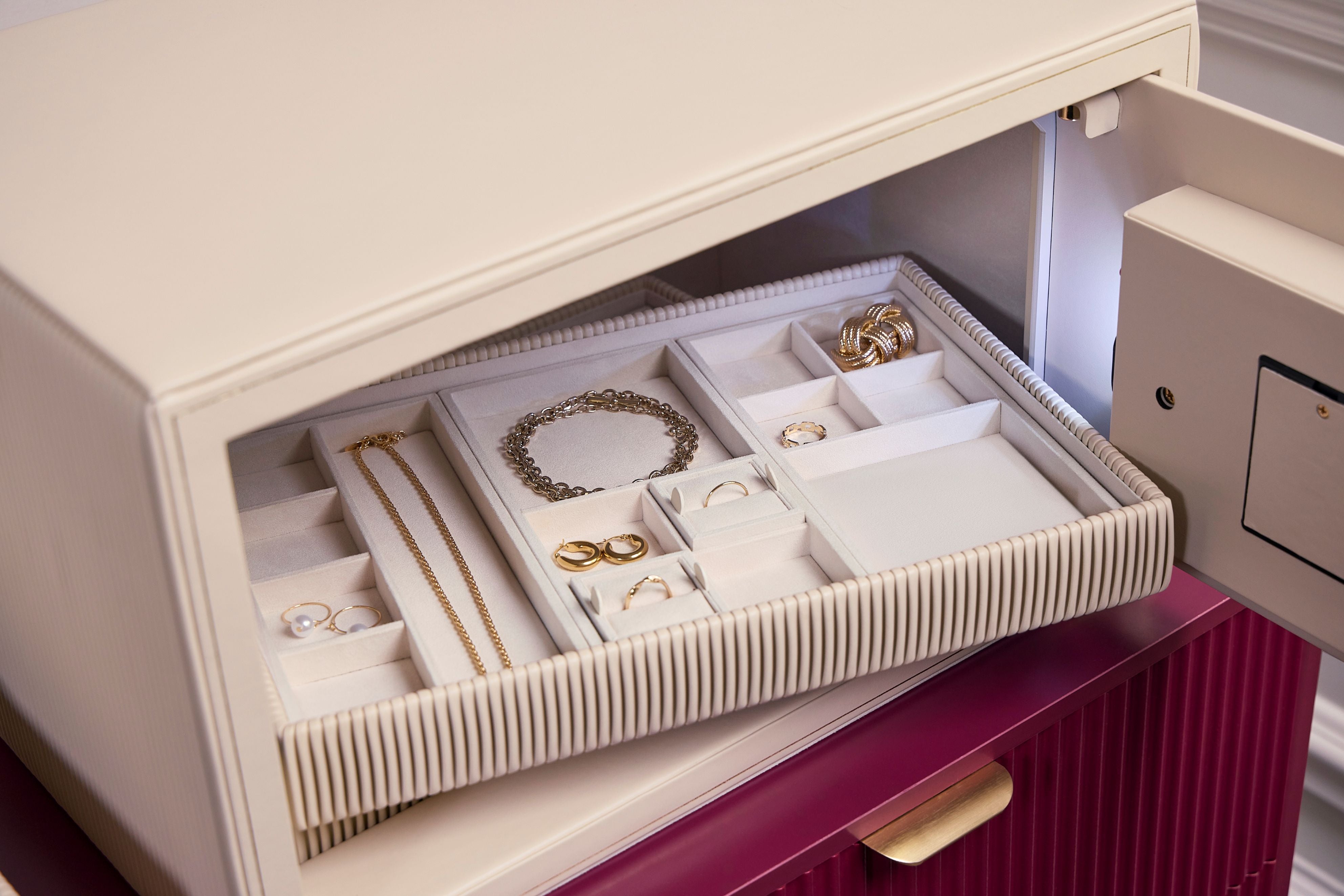 Coffre fort à bijoux Bella Den Wolf 1834 de couleur crème à ouverture biométrique. Vue lifestyle, avec le coffre ouvert posé sur une commode rouge et un zoom sur les plateaux à bijoux remplis de bijoux à l'intérieur