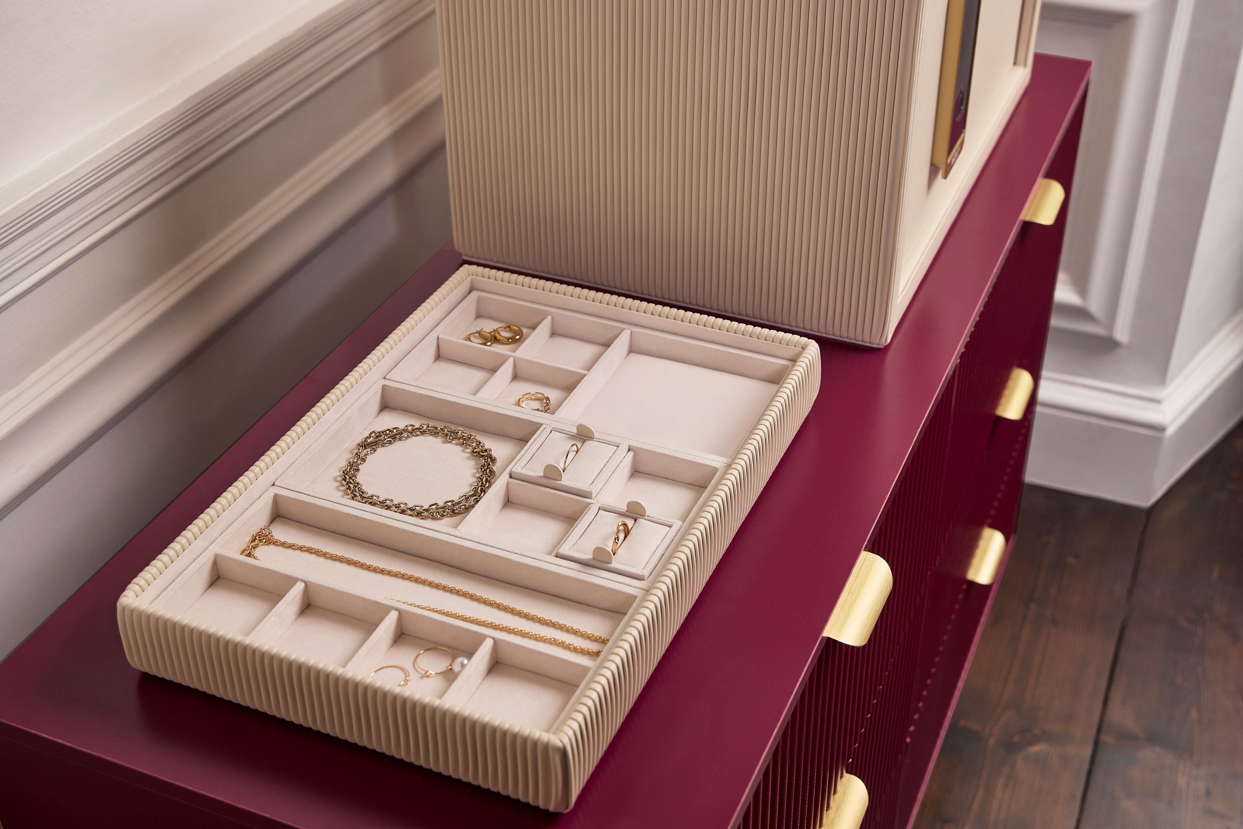 Coffre fort à bijoux Bella Den Wolf 1834 de couleur crème à ouverture biométrique. Vue lifestyle, avec le coffre fermé posé sur une commode rouge et un zoom sur le plateau de rangement à bijoux rempli de bijoux précieux et posé sur la commode à côté