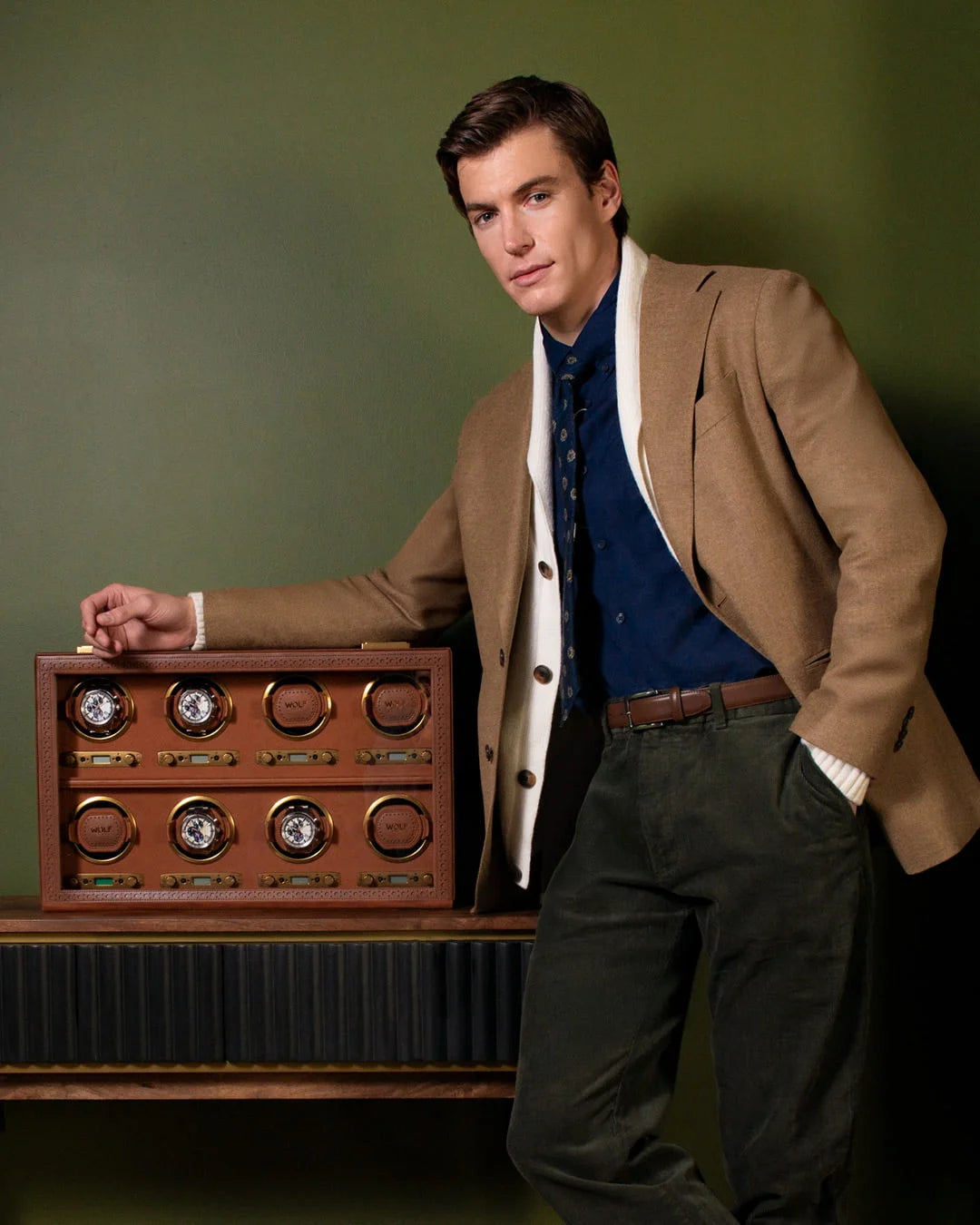 Homme élégant anglais en costume avec une chemise bleu marine et une cravate bleu, accoudé sur un remontoir à montres automatiques Wolf 1834, collection Brogue, avec une esthétique vintage et intemporelle des célèbres chaussures britanniques « brogue » - Arrière plan vert