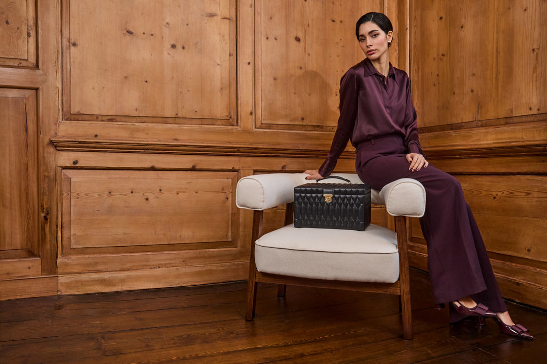 Femme avec une chemise en soie prune se tenant devant une boiserie, assise sur l'accoudoir d'un fauteuil blanc, avec posé sur le fauteuil blanc une boîte à bijoux noire de la collection Lauren de Wolf