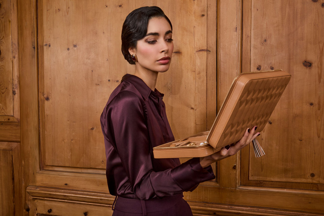 Femme avec une chemise en soie prune se tenant devant une boiserie et regardant sa boîte à bijoux de voyage ouverte de couleur caramel