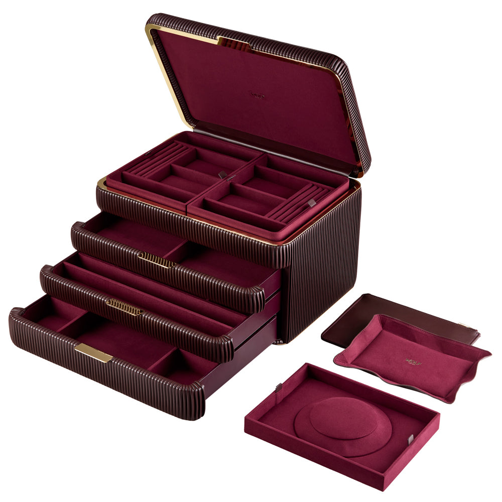 Armoire à bijoux couleur bordeaux de la collection Bella de la manufacture Wolf 1834. Vue de transverse, la boîte à bijoux est ouverte, les tiroirs sont tirés, et les accessoires (pochette à bijoux, plateaux à bijoux, reposoir à collier) sont posés à côté de la boîte