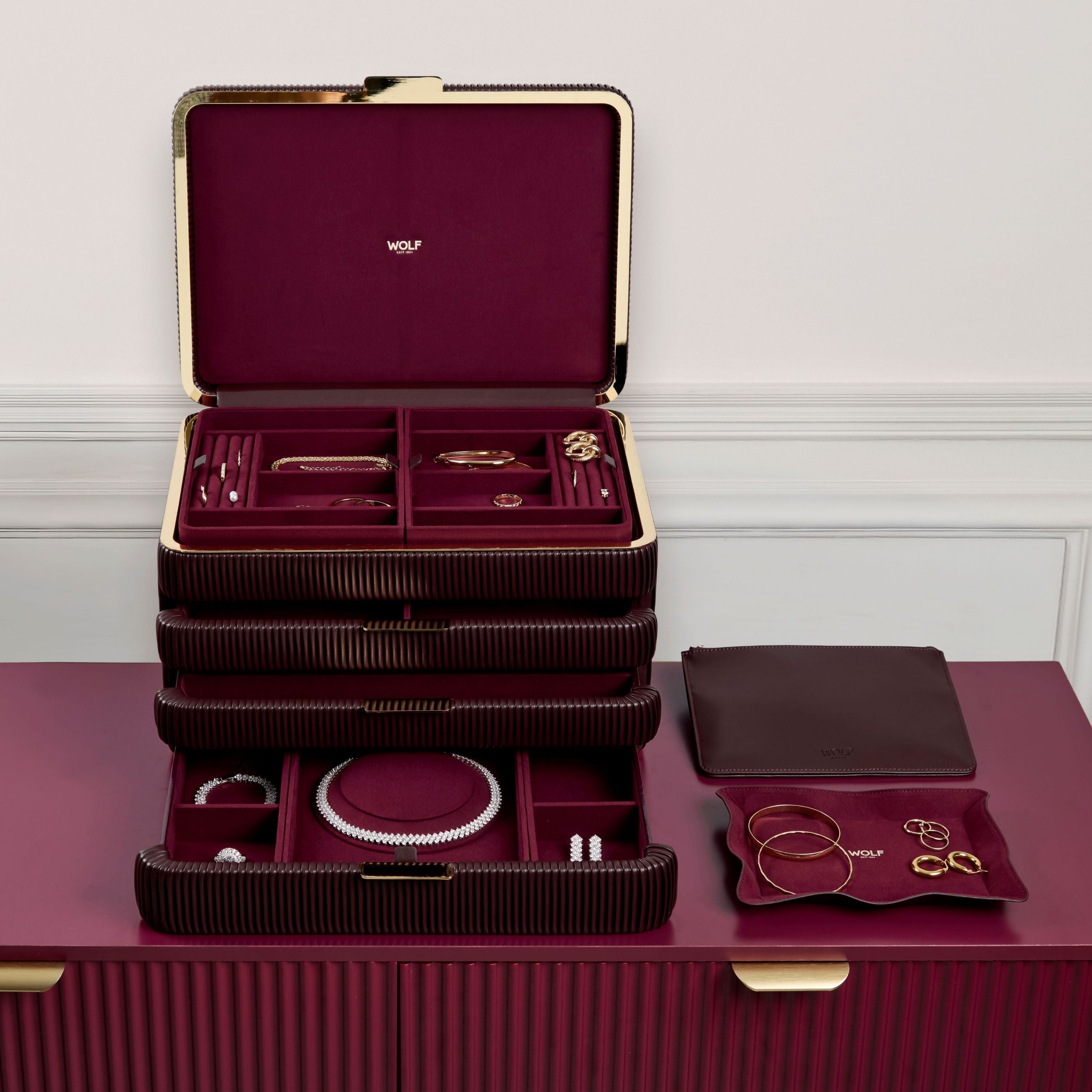 Armoire à bijoux couleur bordeaux de la collection Bella de la manufacture Wolf 1834. Vue de face, posée sur une commode ton sur ton, tiroirs ouverts et remplis de bijoux et accessoires amovibles (plateau à bijoux, pochette à bijoux) posés à côté
