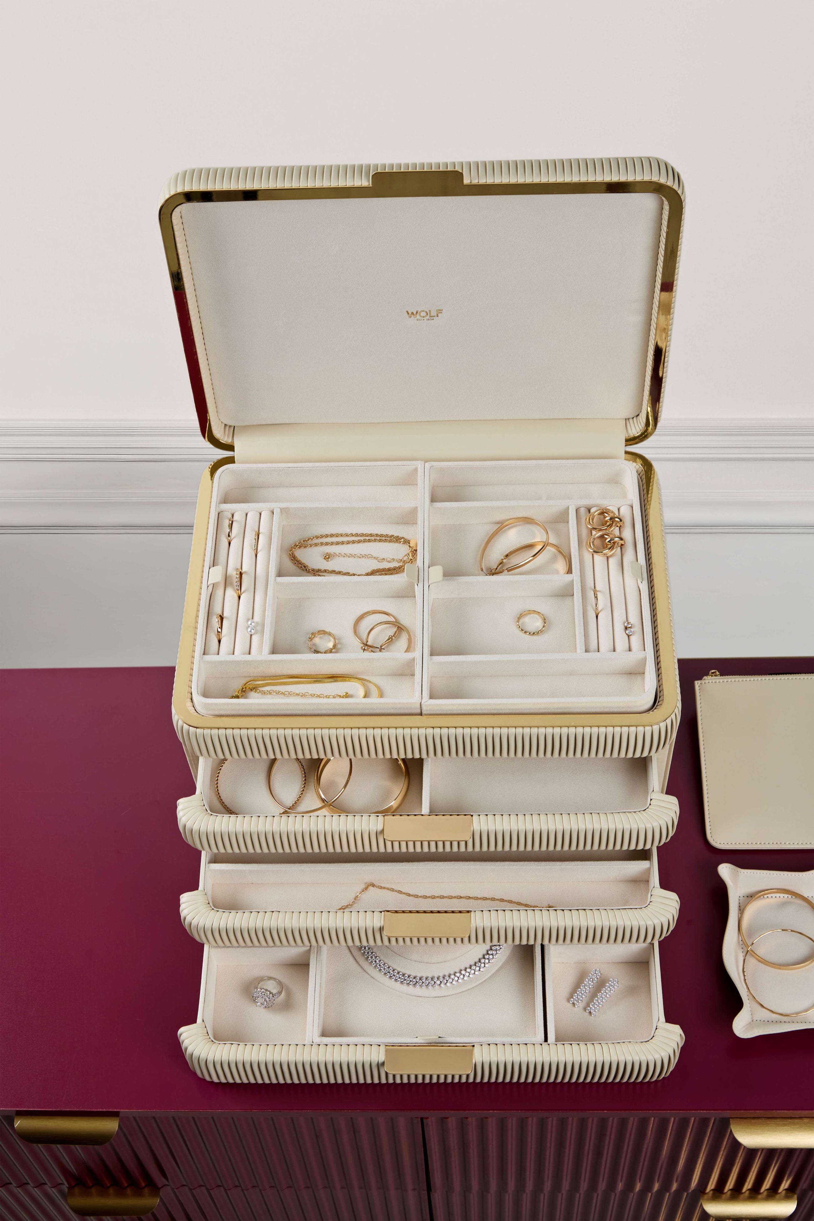 Armoire à bijoux couleur crème de la collection Bella de la manufacture Wolf 1834. Vue de face, posée sur une commode bordeaux devant un mur blanc avec moulure, tiroirs ouverts et remplis de bijoux et accessoires amovibles (plateaux à bijoux, pochette à bijoux) posés à côté