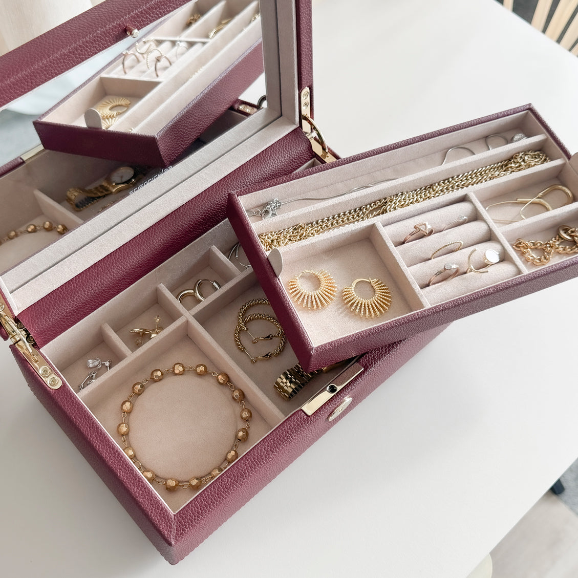 Boîte à bijoux en cuir bordeaux de la collection Vitoria. Vue ouverte avec les compartiments intérieurs sortis et remplis de bijoux. La boîte est posée sur une commode