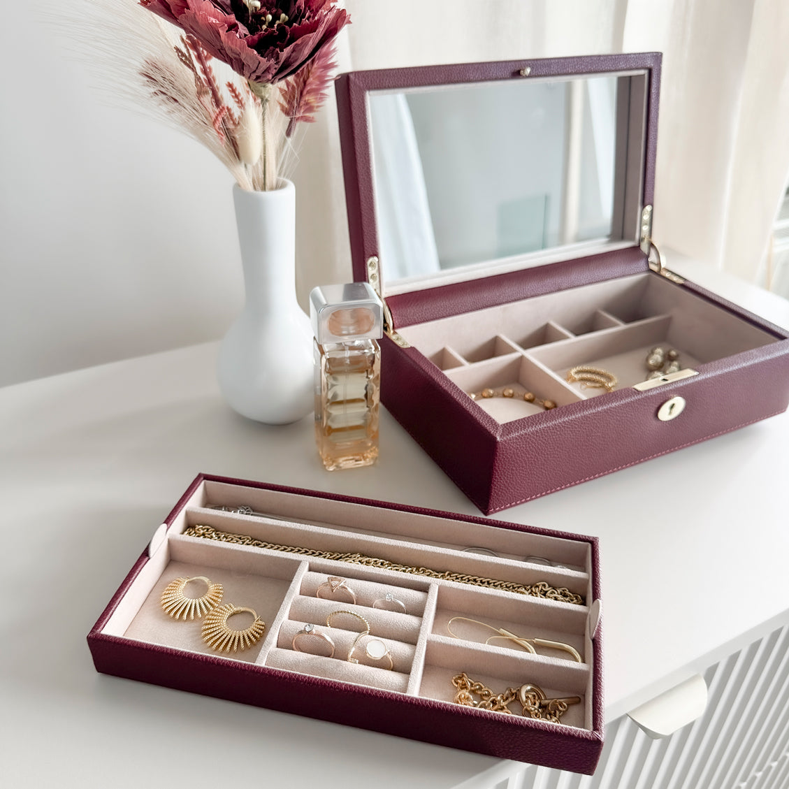 Boîte à bijoux en cuir bordeaux de la collection Vitoria. Vue ouverte remplie de bijoux. La boîte et le compartiment intérieur sont posés sur une commode