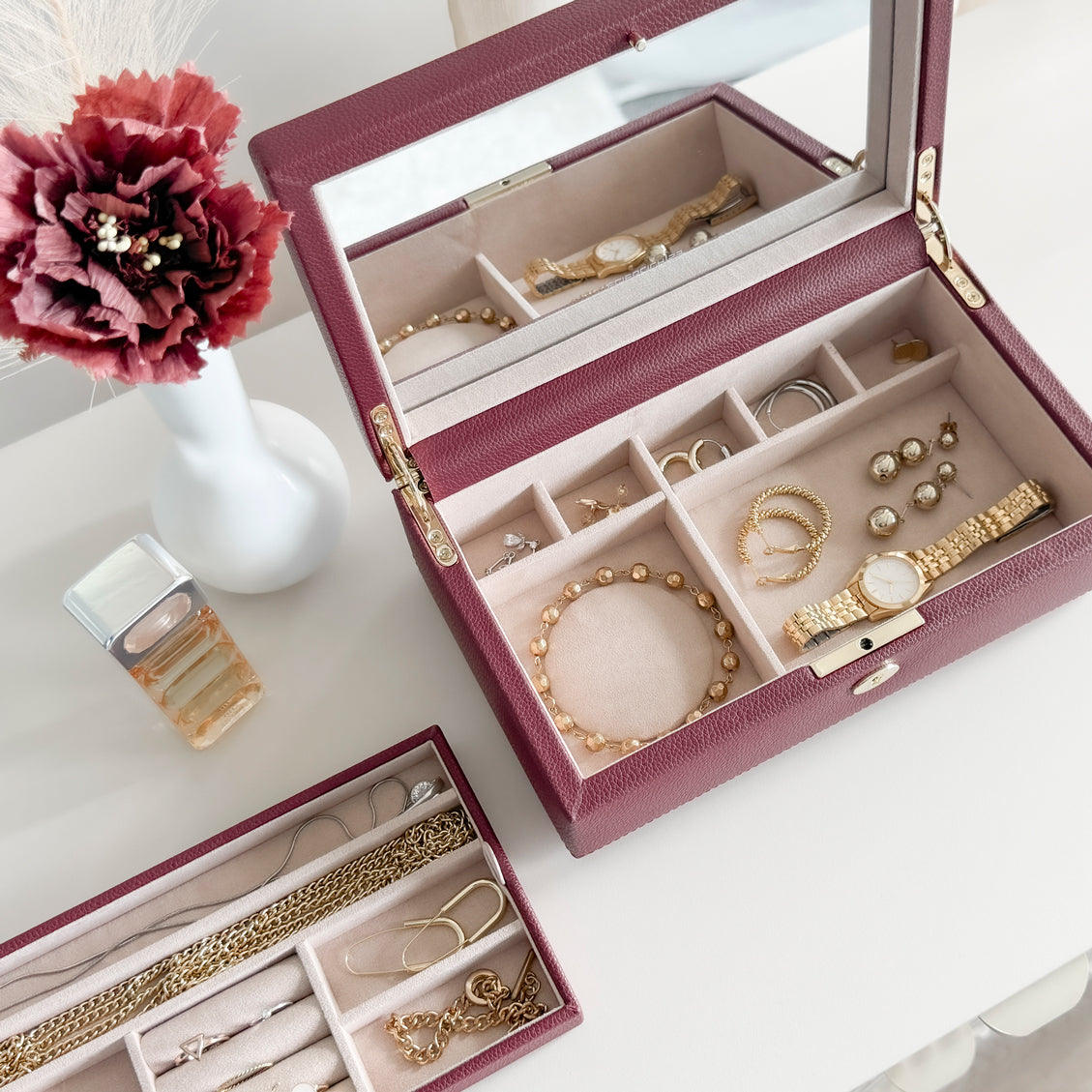Boîte à bijoux en cuir bordeaux de la collection Vitoria. Vue ouverte remplie de bijoux. La boîte et le compartiment intérieur sont posés sur une commode