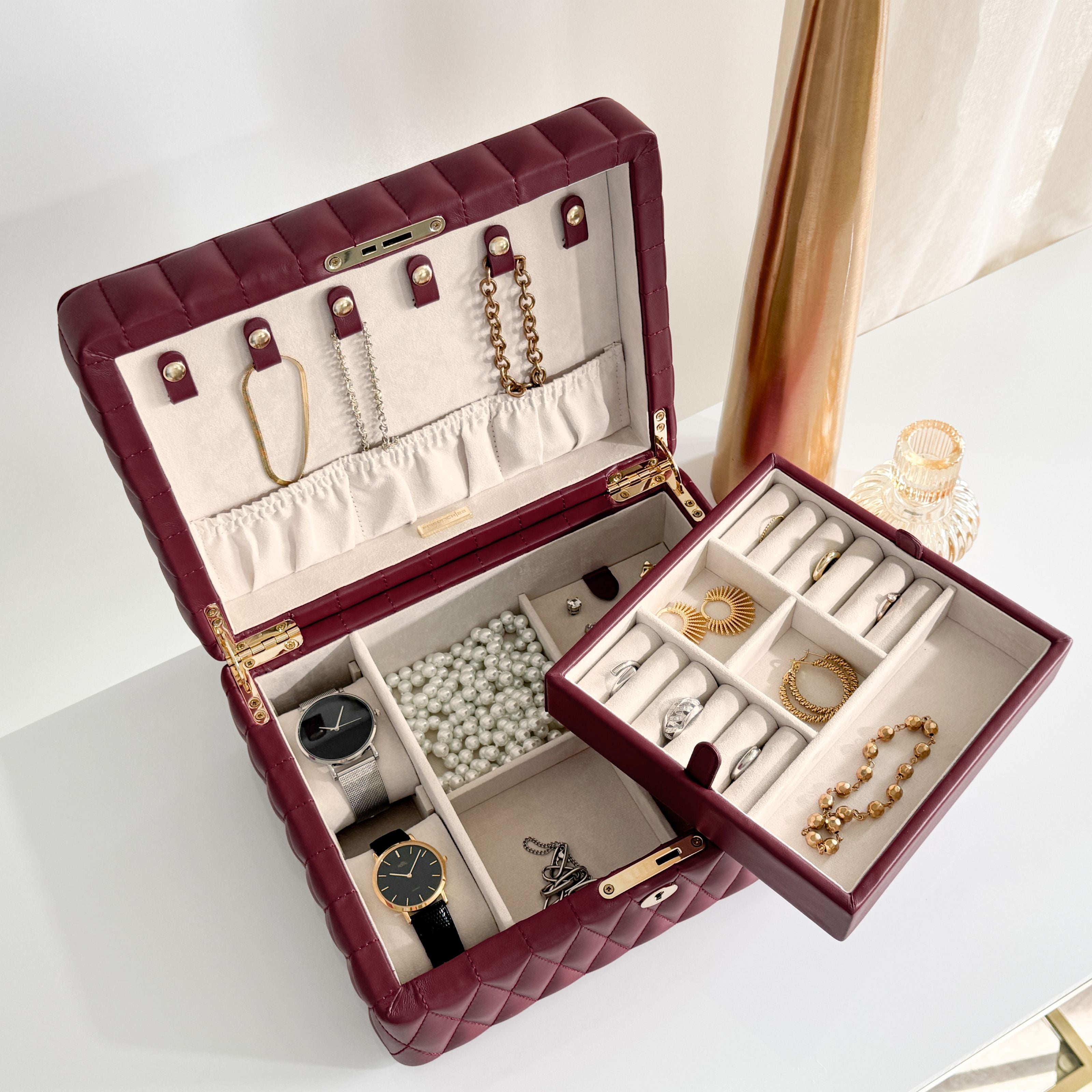 Boîte à bijoux matelassé Saphira bordeaux. La boîte est ouverte, remplie de bijoux et posée sur une surface plane blanche et lisse
