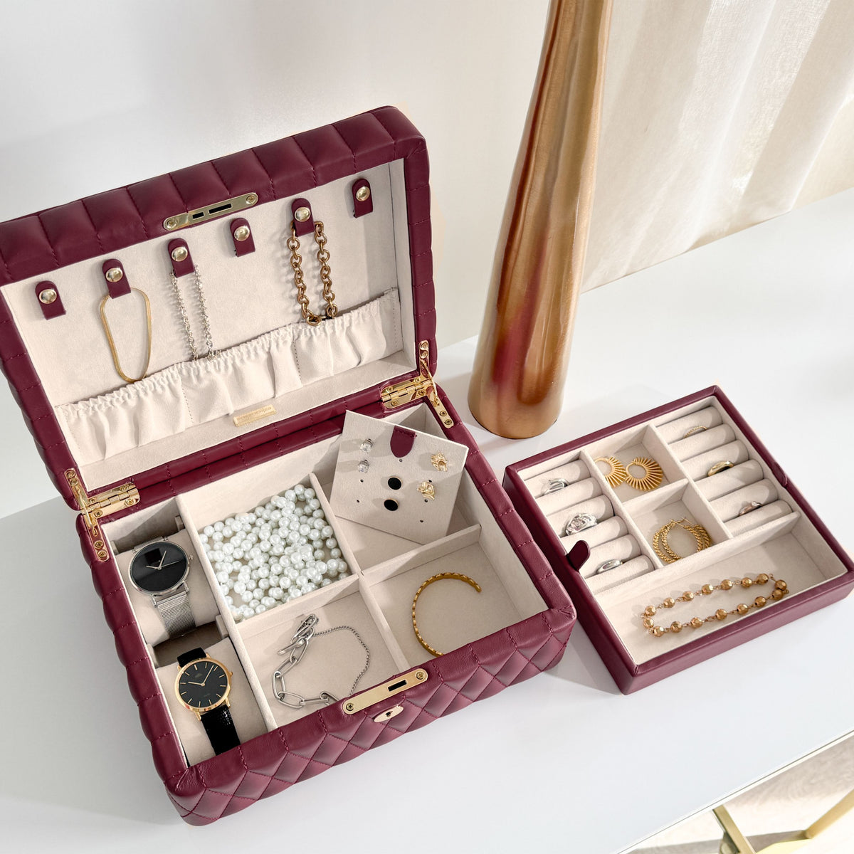 Boîte à bijoux matelassé Saphira bordeaux. Vue de la boîte de haut, la boîte est posée sur une surface plane blanche et est remplie de montres et bijoux dans des teintes dorées