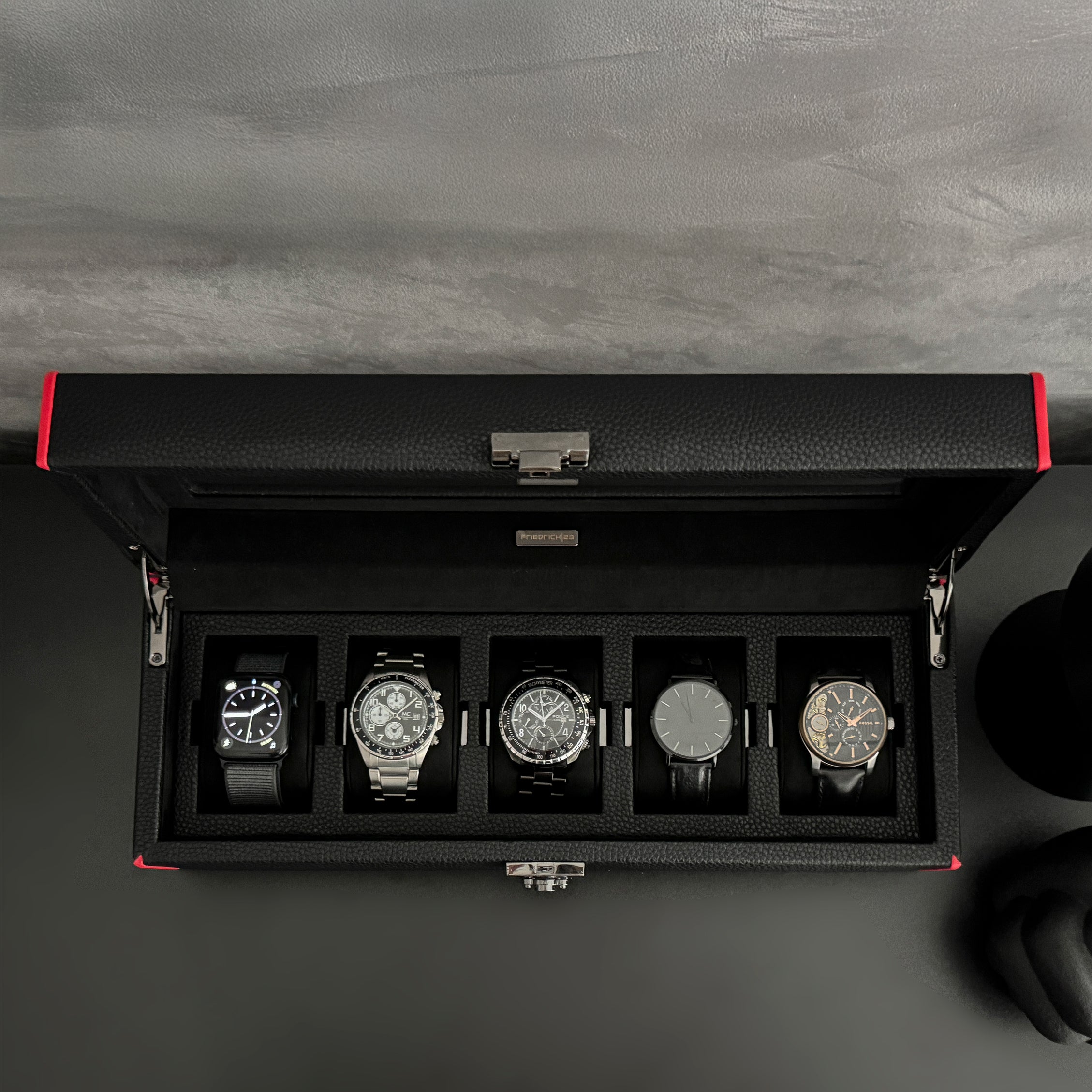 Boîte à montre vitrée en cuir noir avec passepoil rouge Maranello, cinq emplacements. La boîte est posée sur une commode noire. Elle est ouverte, avec une vue de haut, et remplie de montres