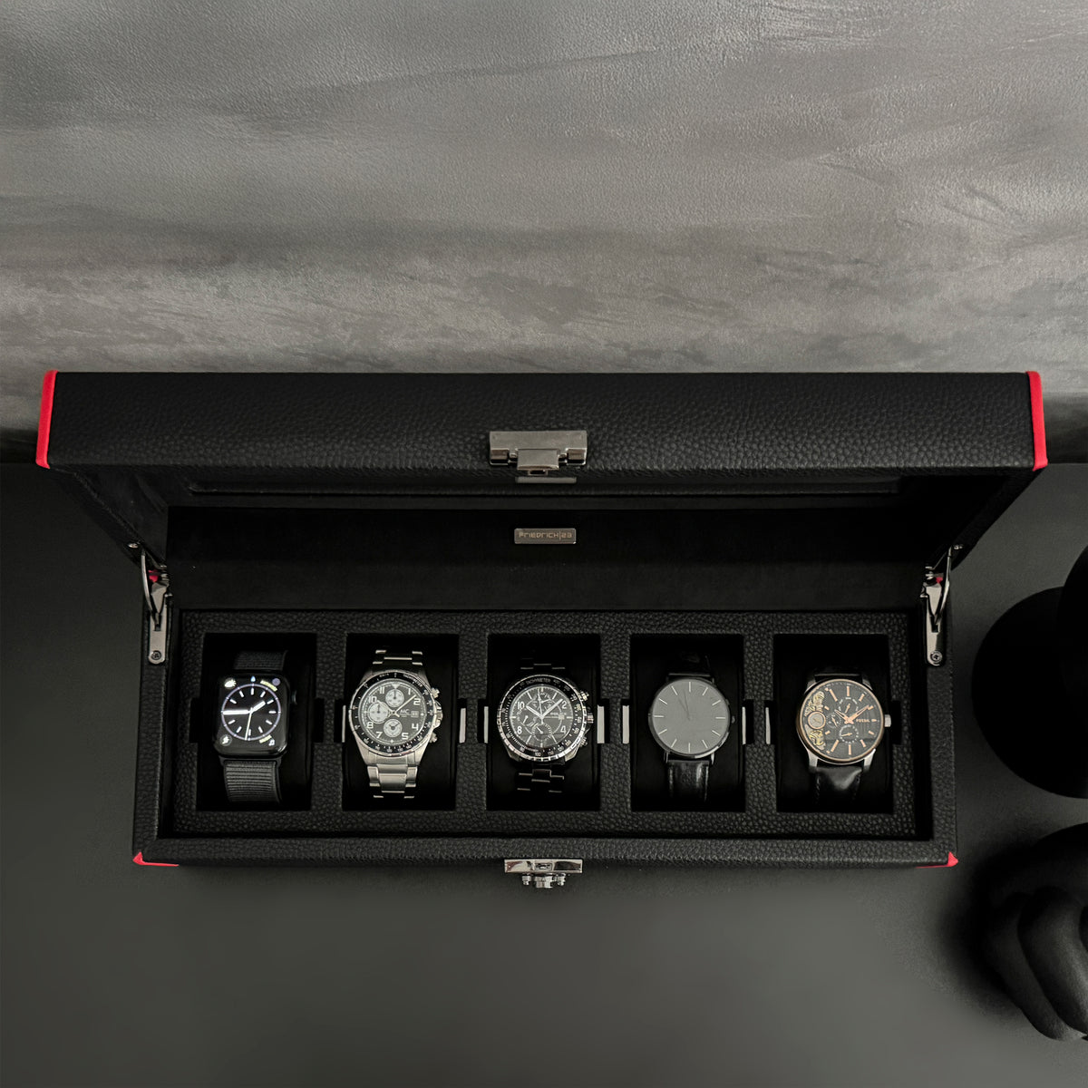 Boîte à montre vitrée en cuir noir avec passepoil rouge Maranello, cinq emplacements. La boîte est posée sur une commode noire. Elle est ouverte, avec une vue de haut, et remplie de montres