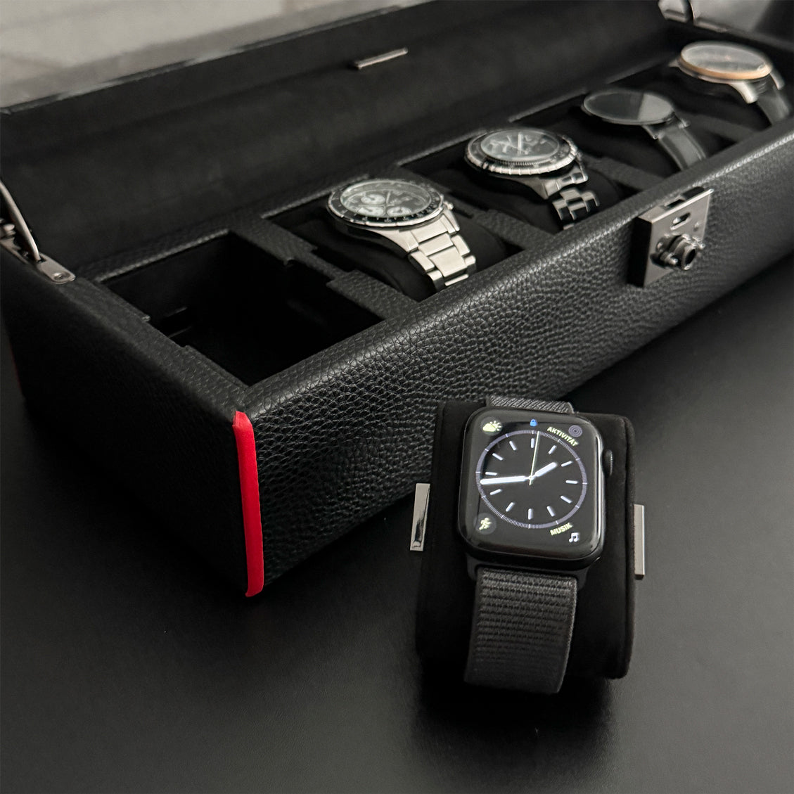 Boîte à montre vitrée en cuir noir avec passepoil rouge Maranello, cinq emplacements. La boîte est posée sur une commode noire. Elle est ouverte, avec une vue transverse, et remplie de montres. Un coussin à montre est posée à coté de la boîte, avec une montre autour