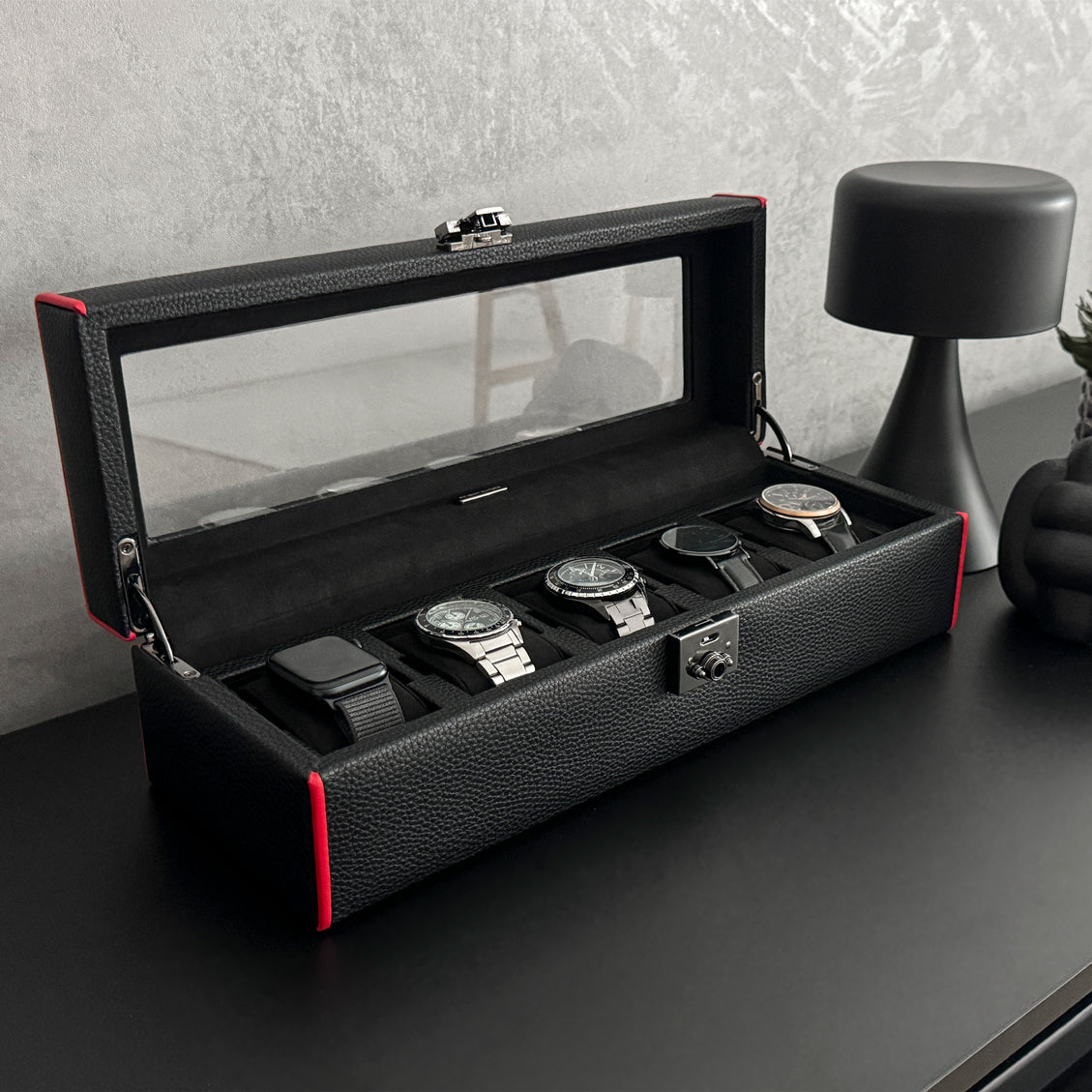 Boîte à montre vitrée en cuir noir avec passepoil rouge Maranello, cinq emplacements. La boîte est posée sur une commode noire. Elle est ouverte, avec une vue transverse, et remplie de montres. 