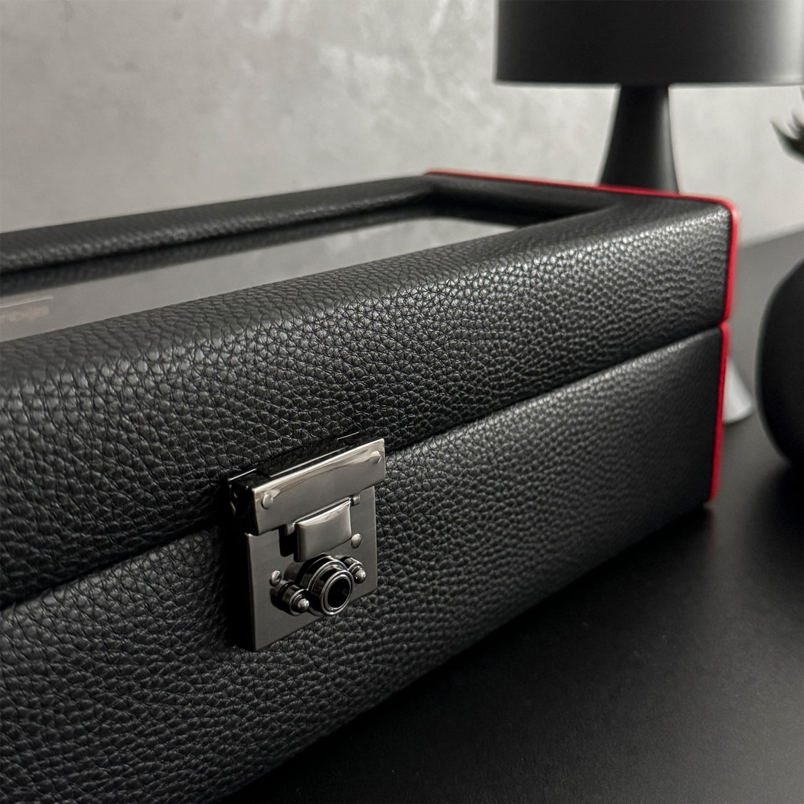Boîte à montre vitrée en cuir noir avec passepoil rouge Maranello, cinq emplacements. La boîte est posée sur une commode noire. Elle est fermée, avec une vue transverse. 