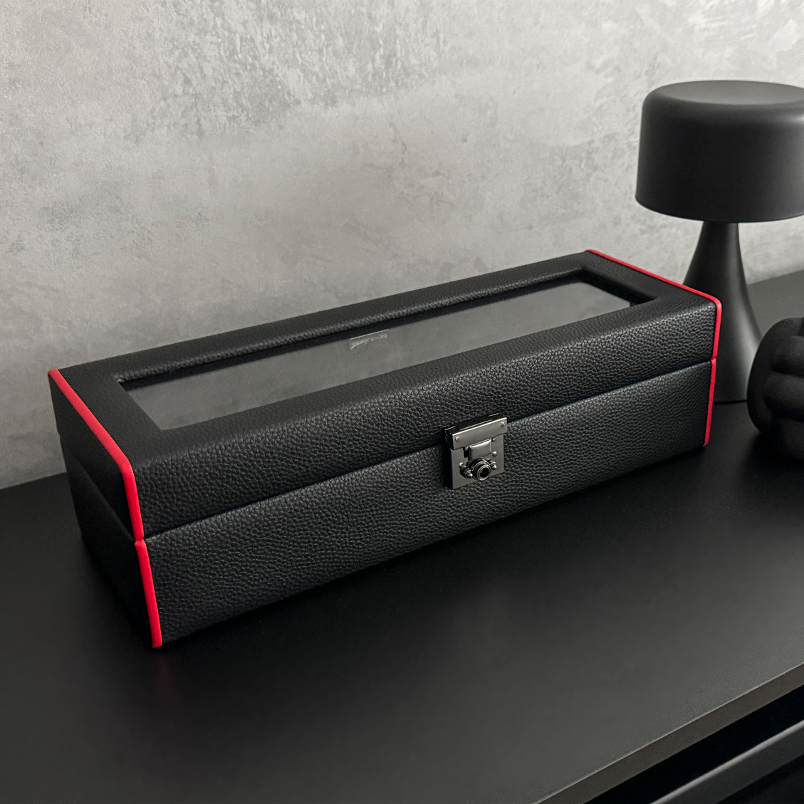 Boîte à montre vitrée en cuir noir avec passepoil rouge Maranello, cinq emplacements. La boîte est posée sur une commode noire. Elle est fermée, avec une vue transverse. 