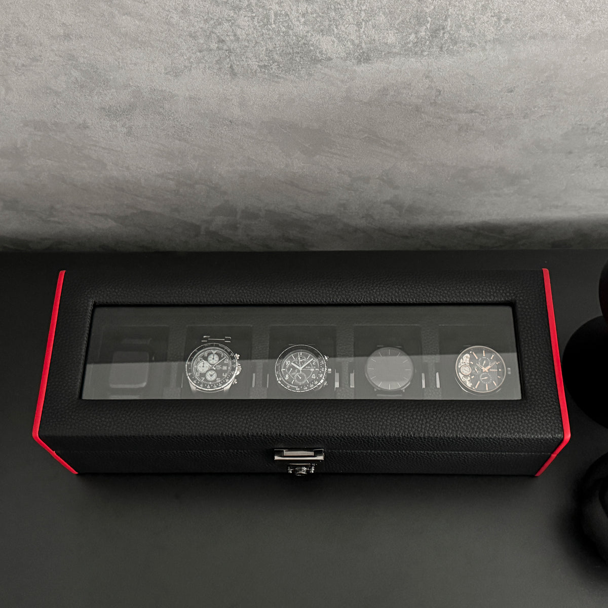 Boîte à montre vitrée en cuir noir avec passepoil rouge Maranello, cinq emplacements. La boîte est posée sur une commode noire. Elle est fermée, remplie de montres, avec une vue de haut.
