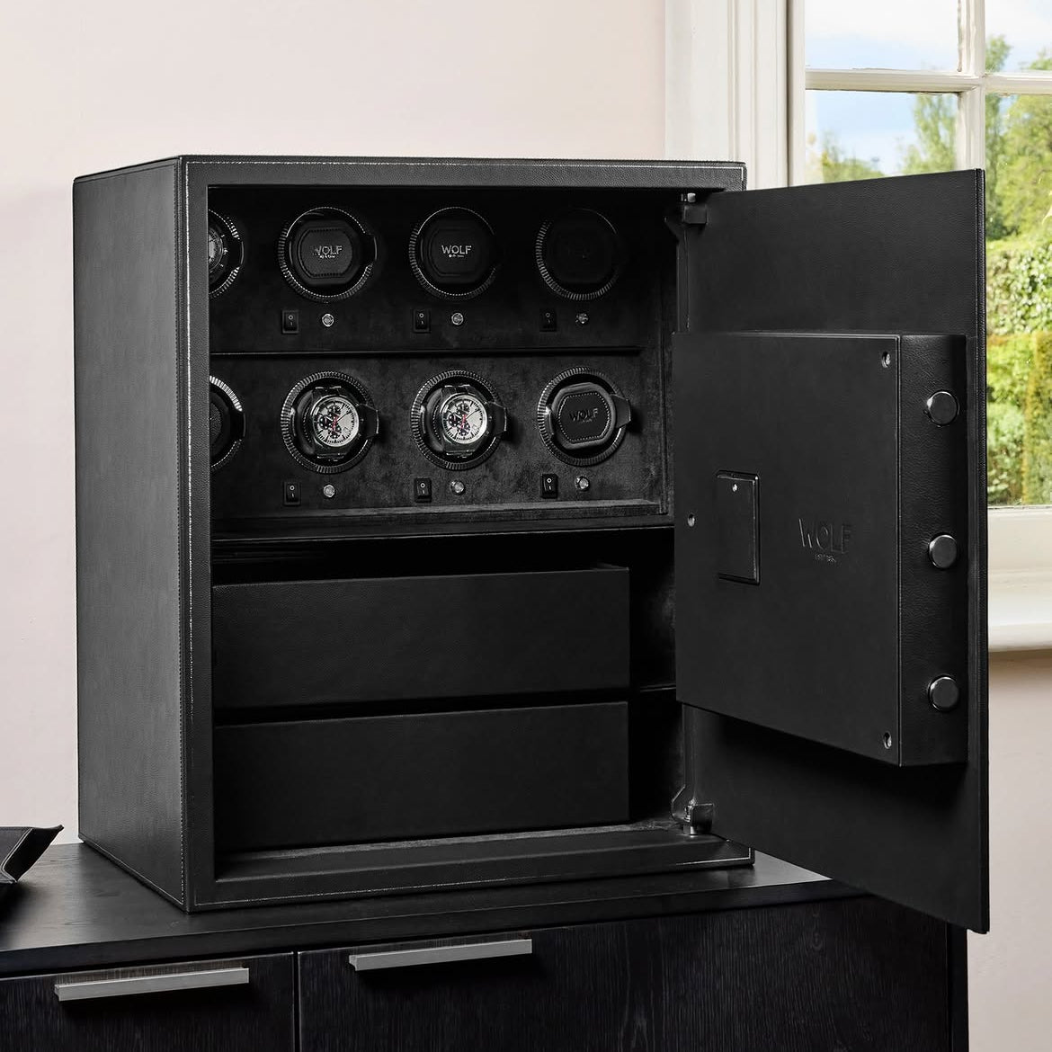Coffre fort biométrique et remontoir huit emplacements de la collection The Den, manufacture Wolf 1834, coloris noir. Le coffre fort est posé sur une commode noire ton sur ton et ouvert. Deux montres sont dans les emplacements remontoirs