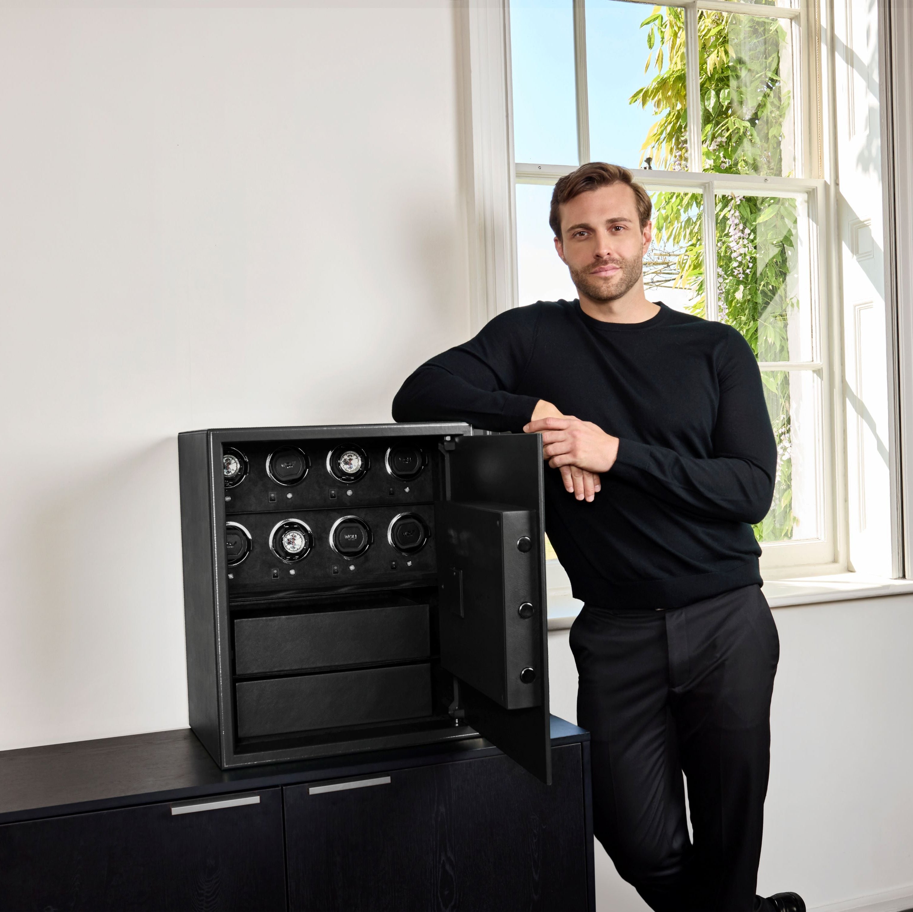 Coffre fort biométrique et remontoir vertemplacements de la collection The Den, manufacture Wolf 1834, coloris noir.
Le coffre fort est posé sur une commode noir et un homme est debout sur la gauche du coffre, accoudé dessus, les mains l'une sur l'autre