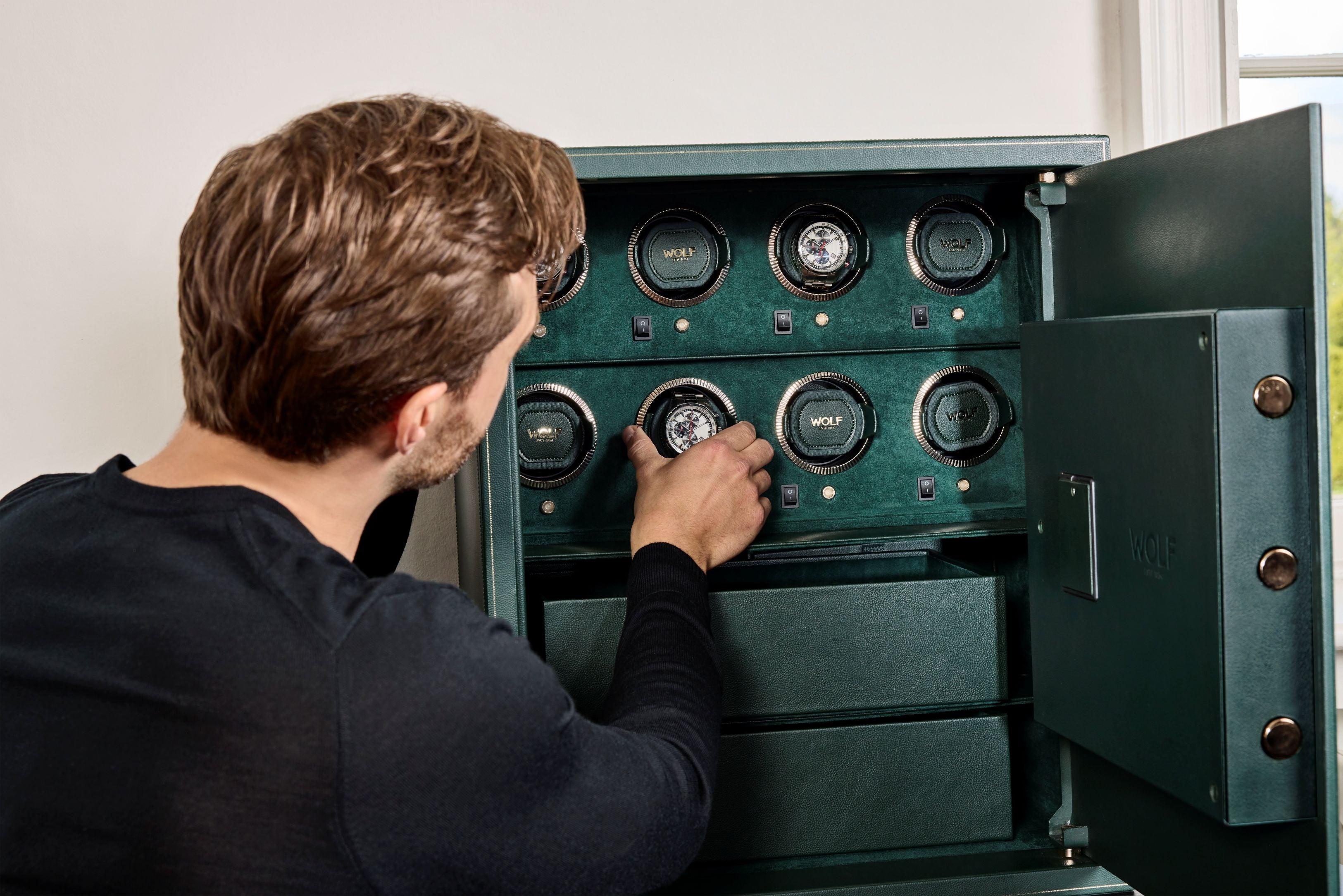 Coffre fort biométrique et remontoir vertemplacements de la collection The Den, manufacture Wolf 1834, coloris vert.
Le coffre fort est ouvert et un homme de dos se saisit d'un emplacement de montre avec une montre sur le dessus pour l'extraire du coffre