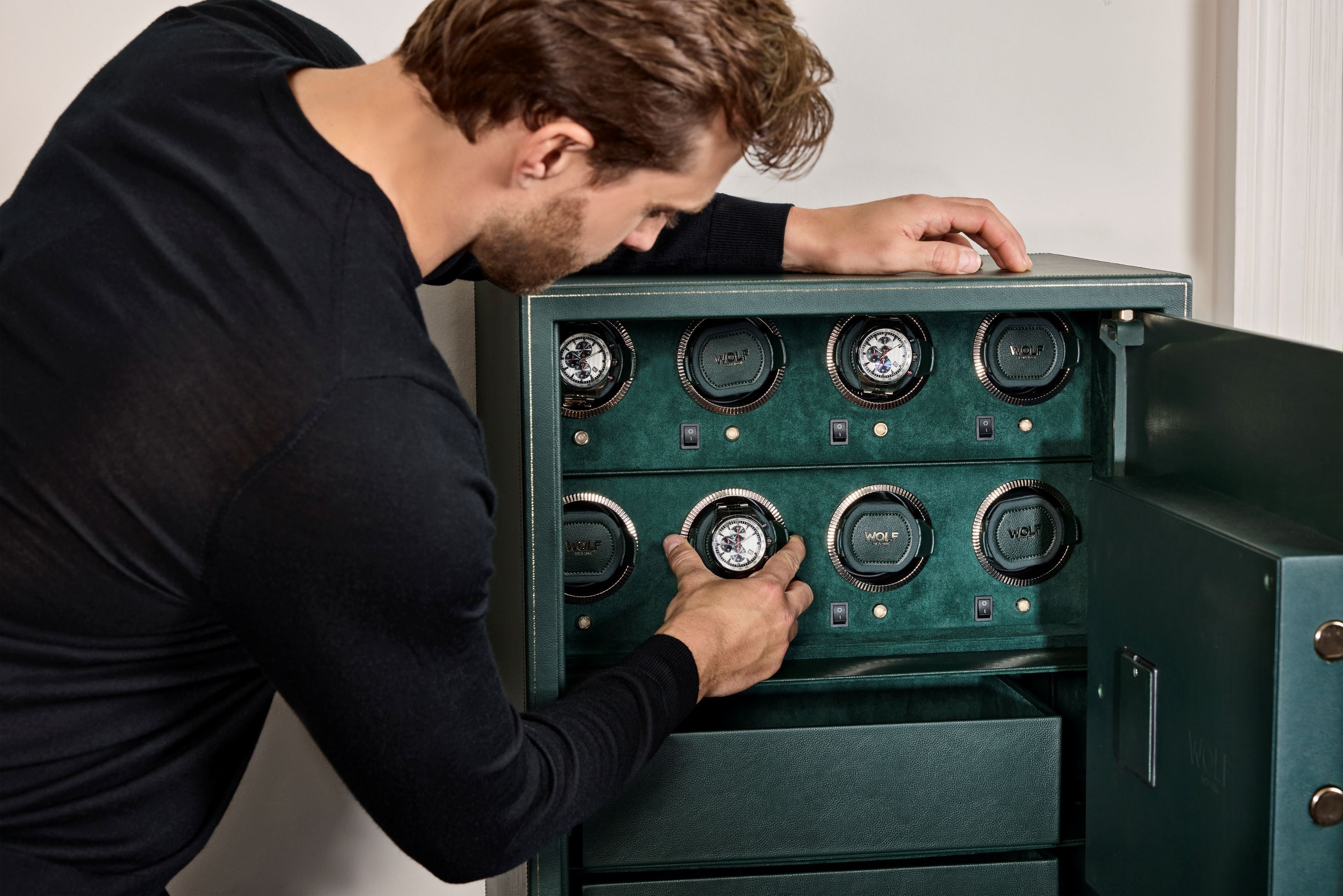 Coffre fort biométrique et remontoir vertemplacements de la collection The Den, manufacture Wolf 1834, coloris vert.
Le coffre fort est ouvert et un homme de dos, accoudé au coffre, se saisit d'un emplacement de montre avec une montre sur le dessus pour l'extraire du coffre