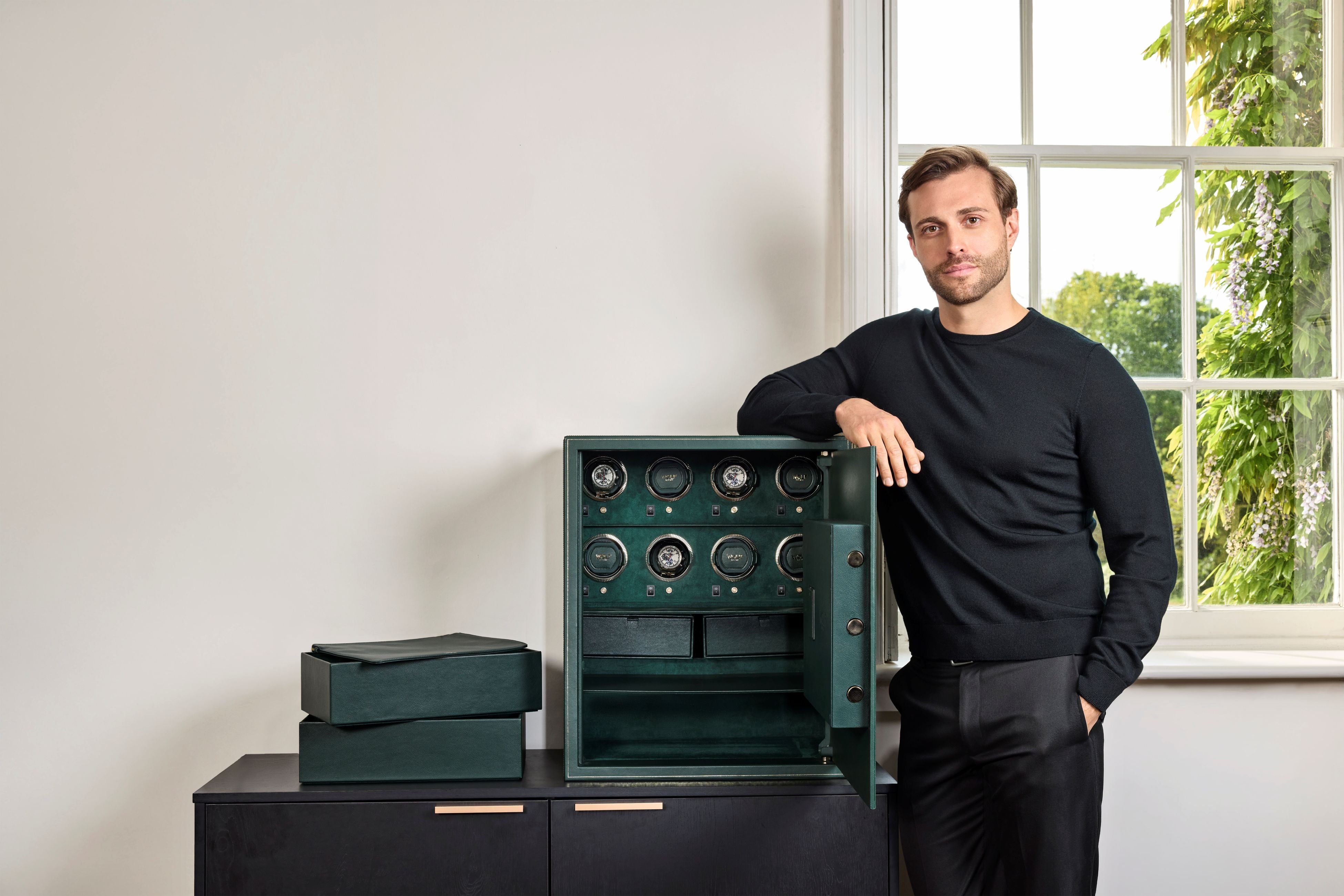 Coffre fort biométrique et remontoir vertemplacements de la collection The Den, manufacture Wolf 1834, coloris vert.
Le coffre fort est ouvert et posé sur une commode noire. Un homme est de face, accoudé sur le coffre, et a sorti les rangements de l'intérieur du coffre