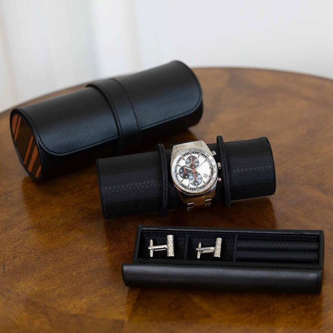 Étui de voyage noir ouvert avec une montre posée sur le coussin cylindrique et un compartiment séparé contenant des boutons de manchette