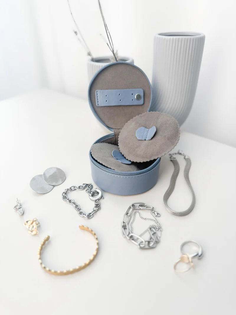 Mini boite à bijoux ronde en cuir véritable Sienna, bleu clair, vue ouverte, posée sur une table à coté d'un vase blanc et entourée de bijoux