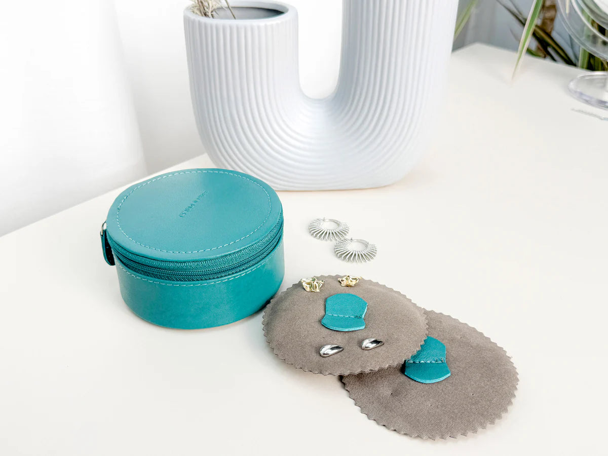 Mini boite à bijoux ronde en cuir véritable Sienna, vert turquoise, vue fermée, posée sur une table avec un vase blanc en fond et des boucles d'oreille sur le côté, et les deux petits coussins à boucle d'oreille sur le côté
