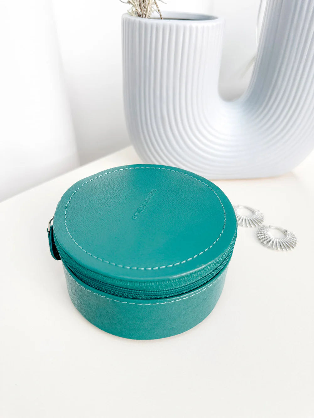 Mini boite à bijoux ronde en cuir véritable Sienna, vert turquoise, vue fermée, posée sur une table avec un vase blanc en fond et des boucles d'oreille sur le côté