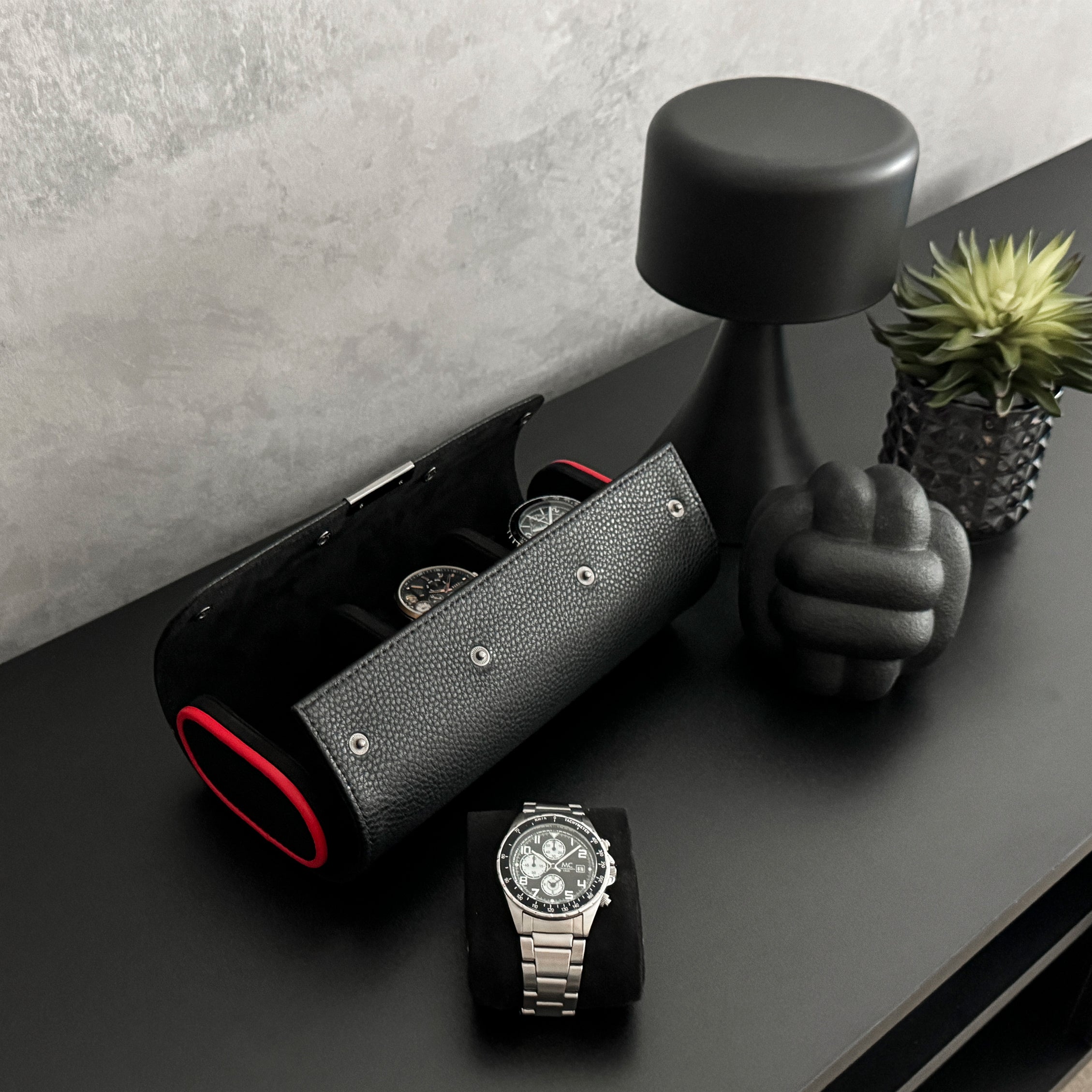 Etui à montre en cuir noir avec passepoil rouge Maranello, trois emplacements. L'étui à montre est posé sur une commode noir, un coussin avec une montre est posé à coté de l'étui. La vue est de haut