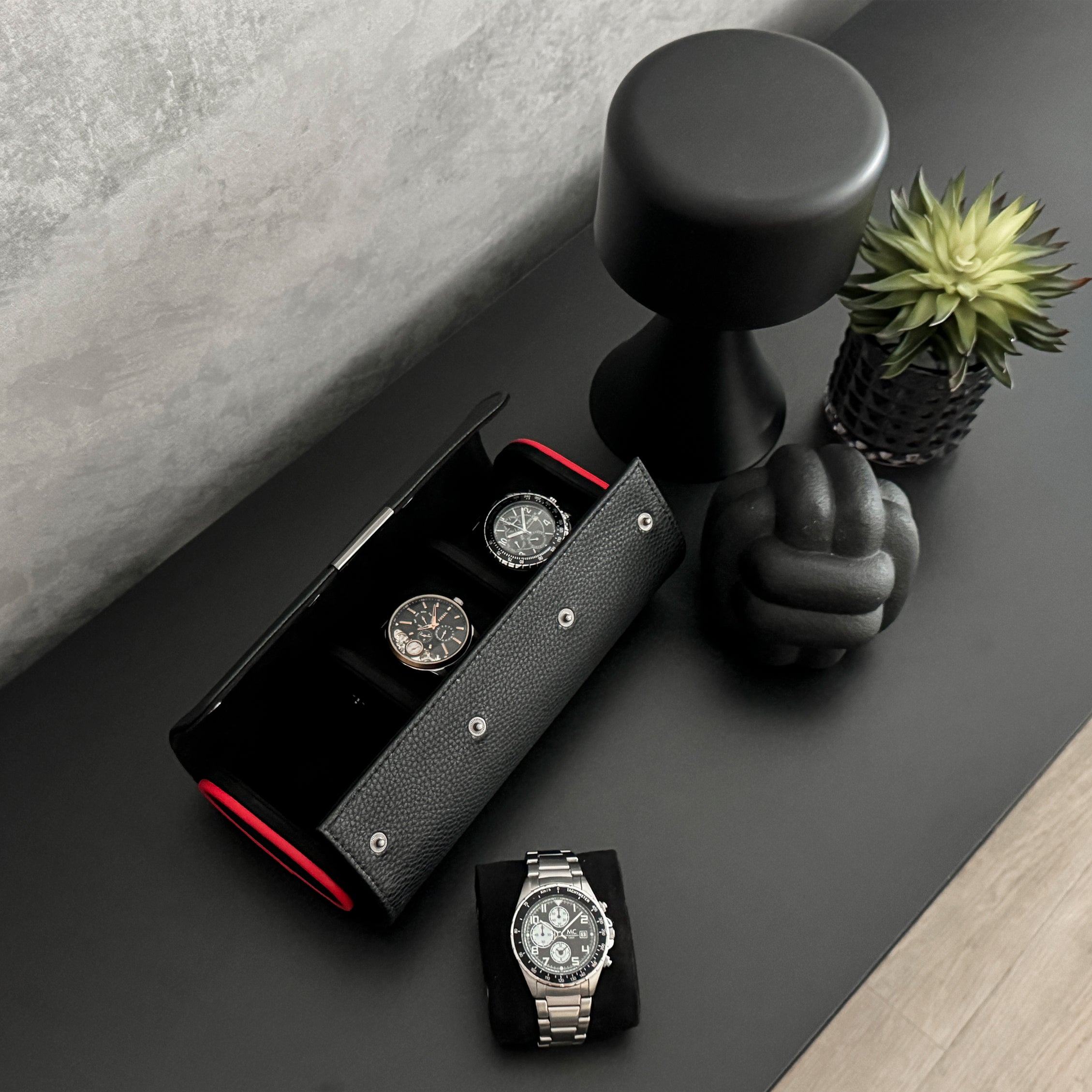 Etui à montre en cuir noir avec passepoil rouge Maranello, trois emplacements. L'étui à montre est posé sur une commode noir, un coussin avec une montre est posé à coté de l'étui. La vue est de haut