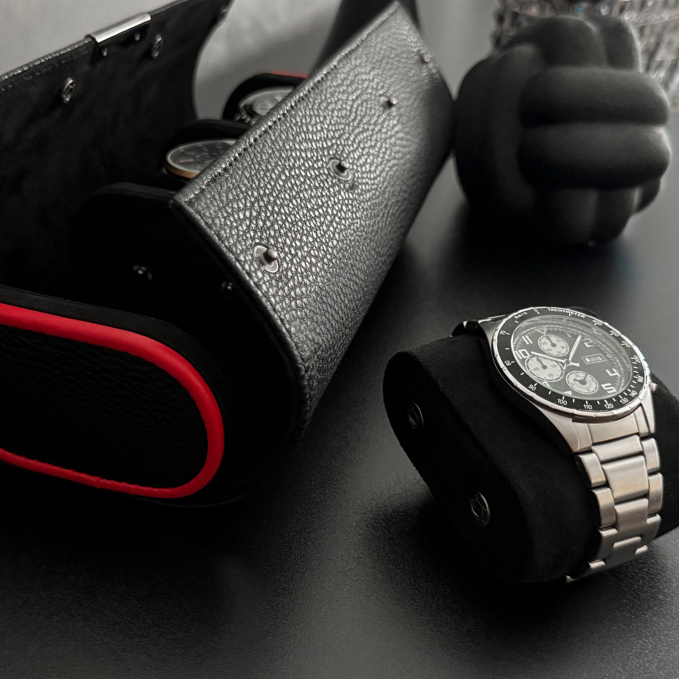 Etui à montre en cuir noir avec passepoil rouge Maranello, trois emplacements. L'étui à montre est posé sur une commode noir, un coussin avec une montre est posé à coté de l'étui