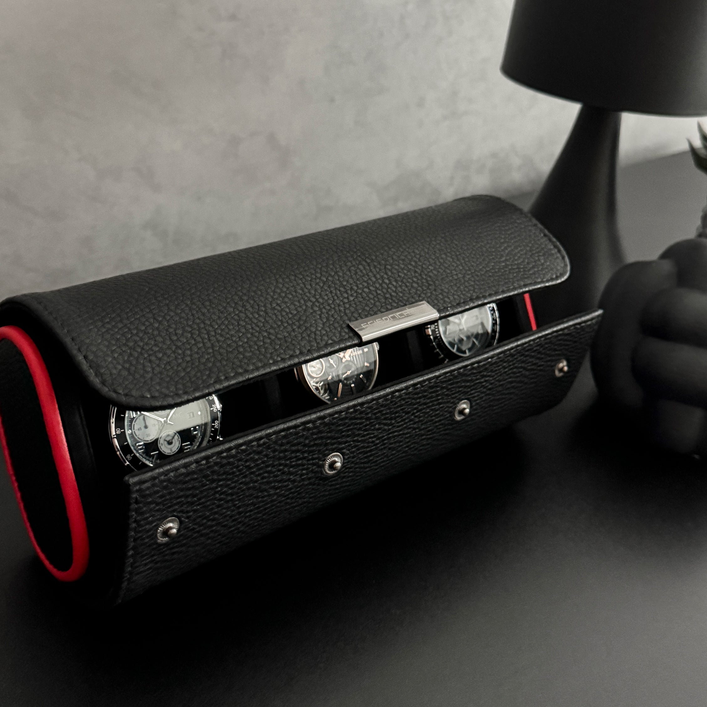 Etui à montre en cuir noir avec passepoil rouge Maranello, trois emplacements. L'étui à montre est posé sur une commode noir, légèrement ouvert et rempli de montres