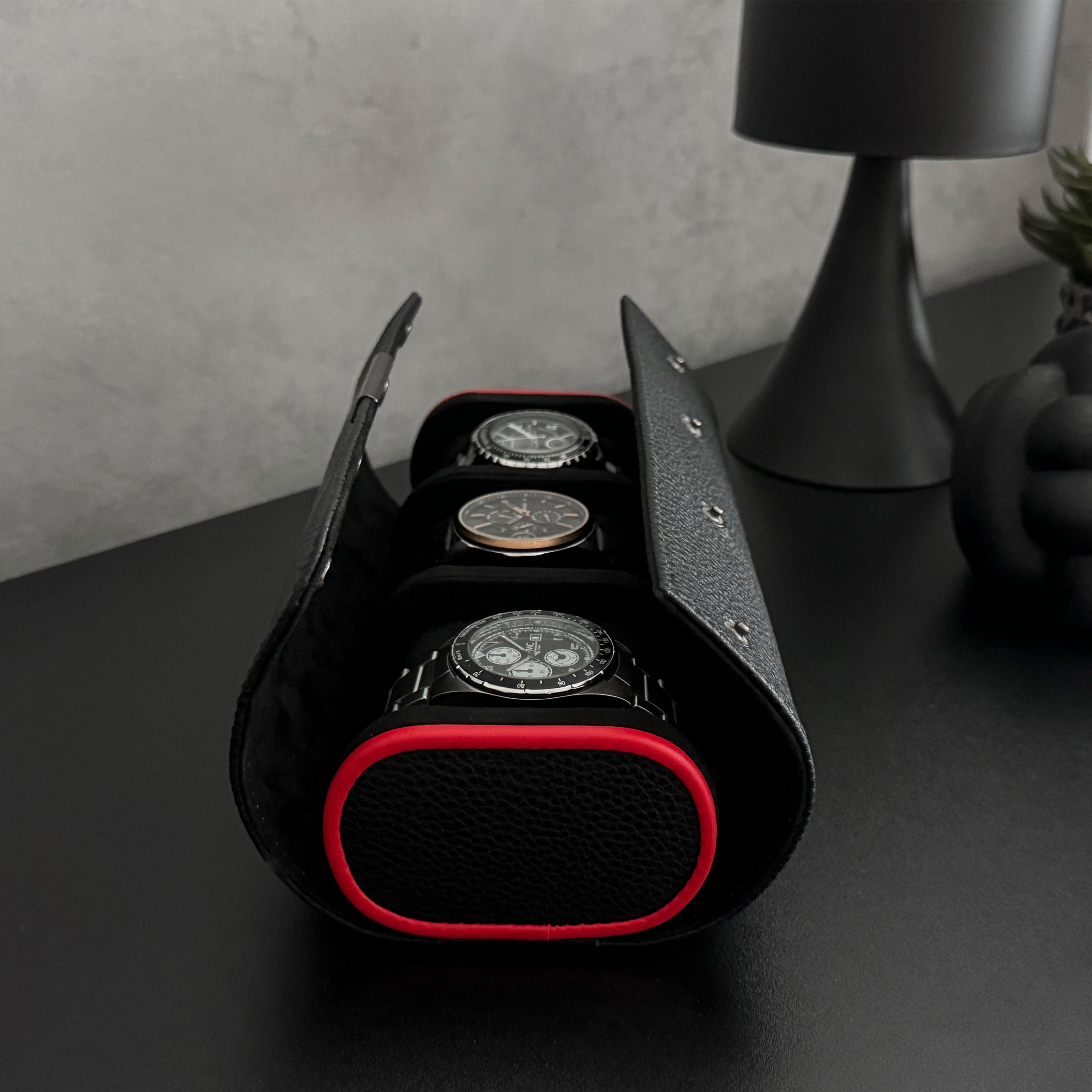Etui à montre en cuir noir avec passepoil rouge Maranello, trois emplacements. L'étui à montre est posé sur une commode noir, ouvert et rempli de montres