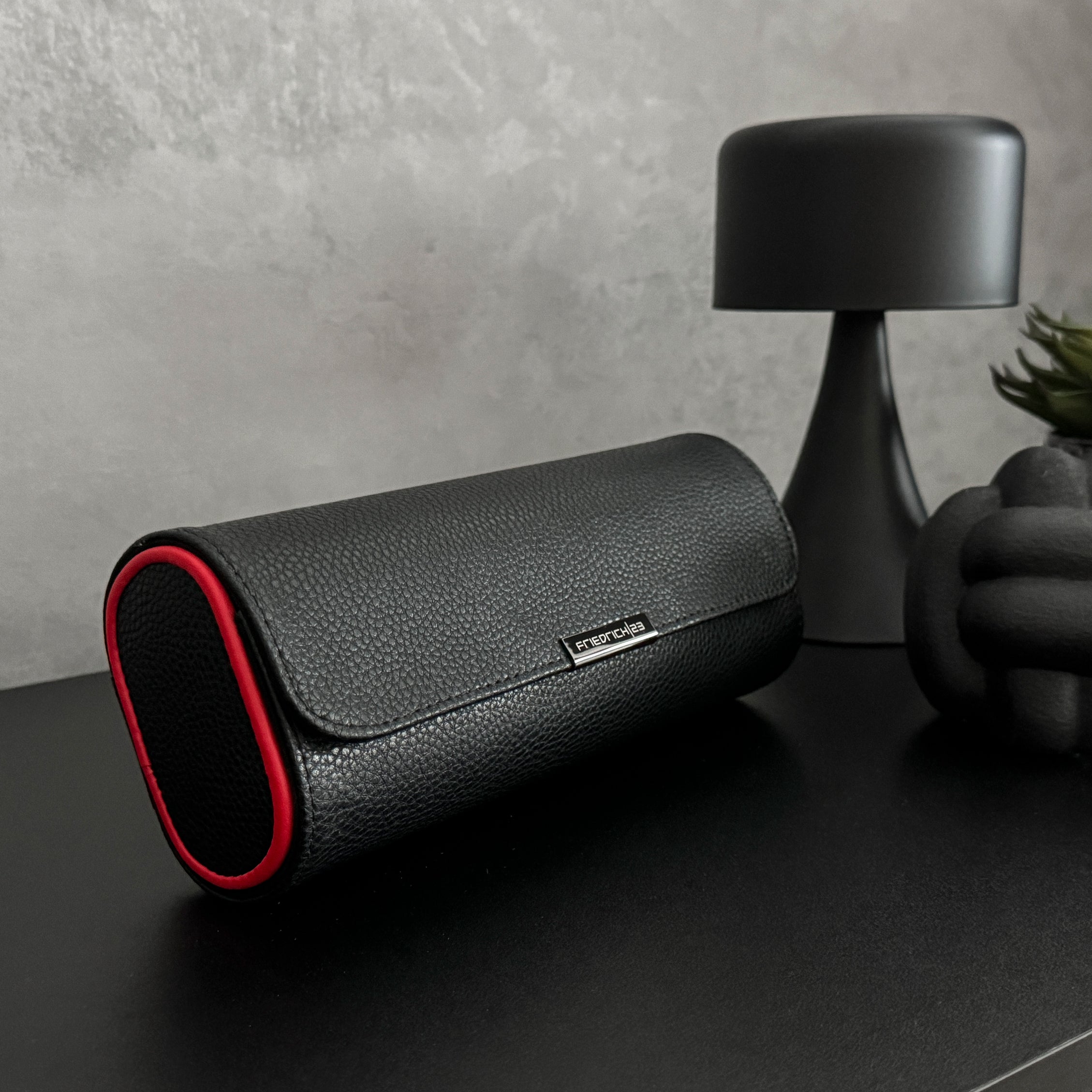 Etui à montre en cuir noir avec passepoil rouge Maranello, trois emplacements. L'étui à montre est posé sur une commode noir, fermé, avec une vue transverse face
